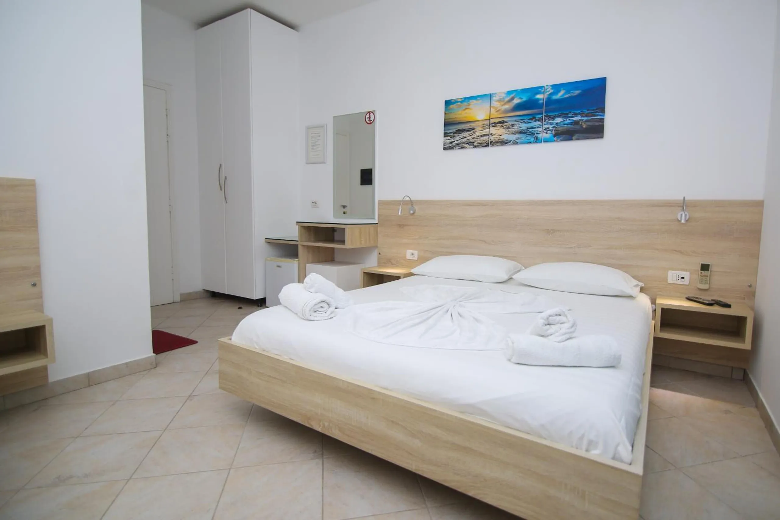 A white double room with a pale-oak bed, towels folded on the white linen and a sea print above the headboard