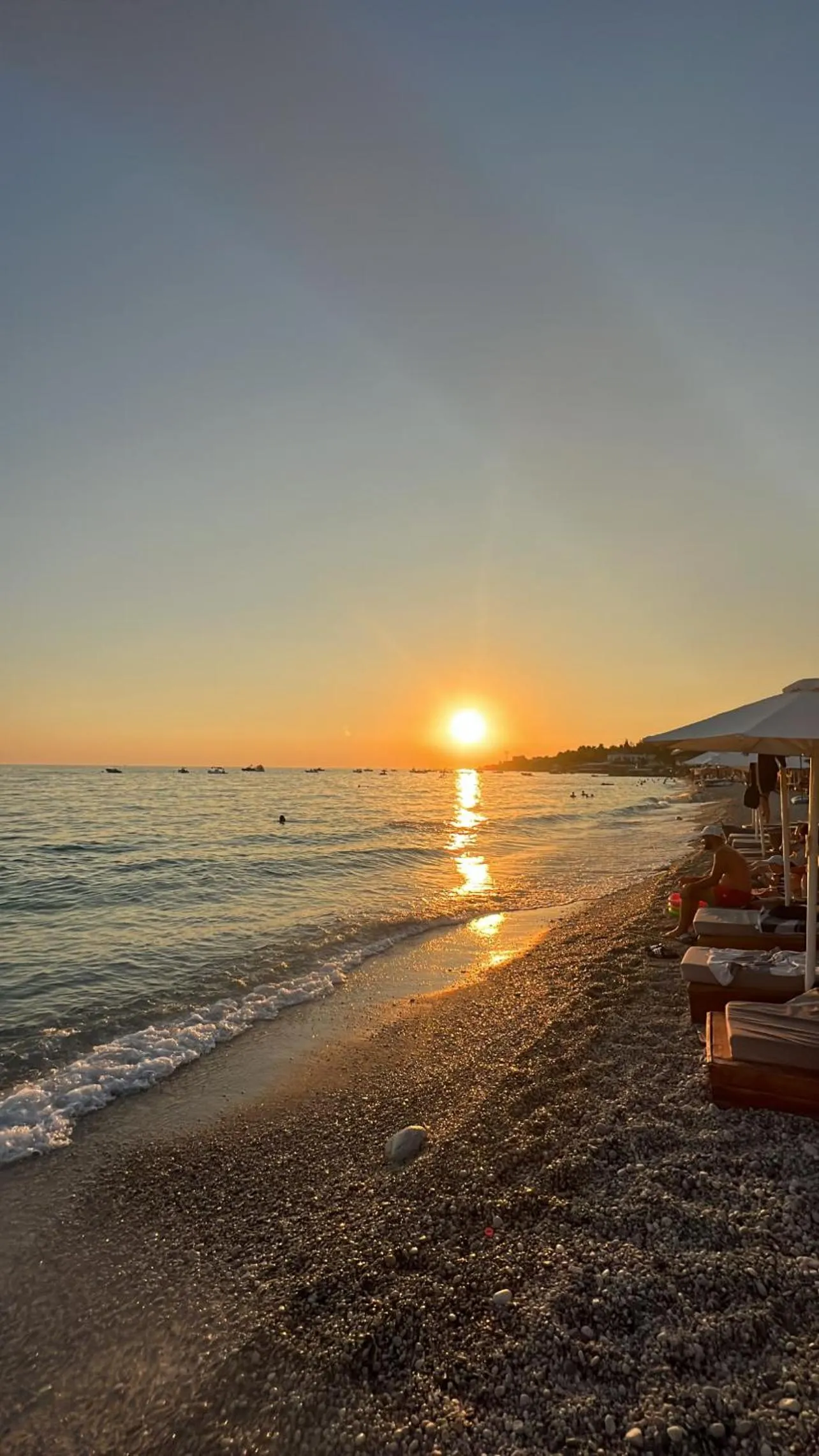 The sun setting into the sea off Dhërmi beach, a gold path on the water and umbrellas along the shore