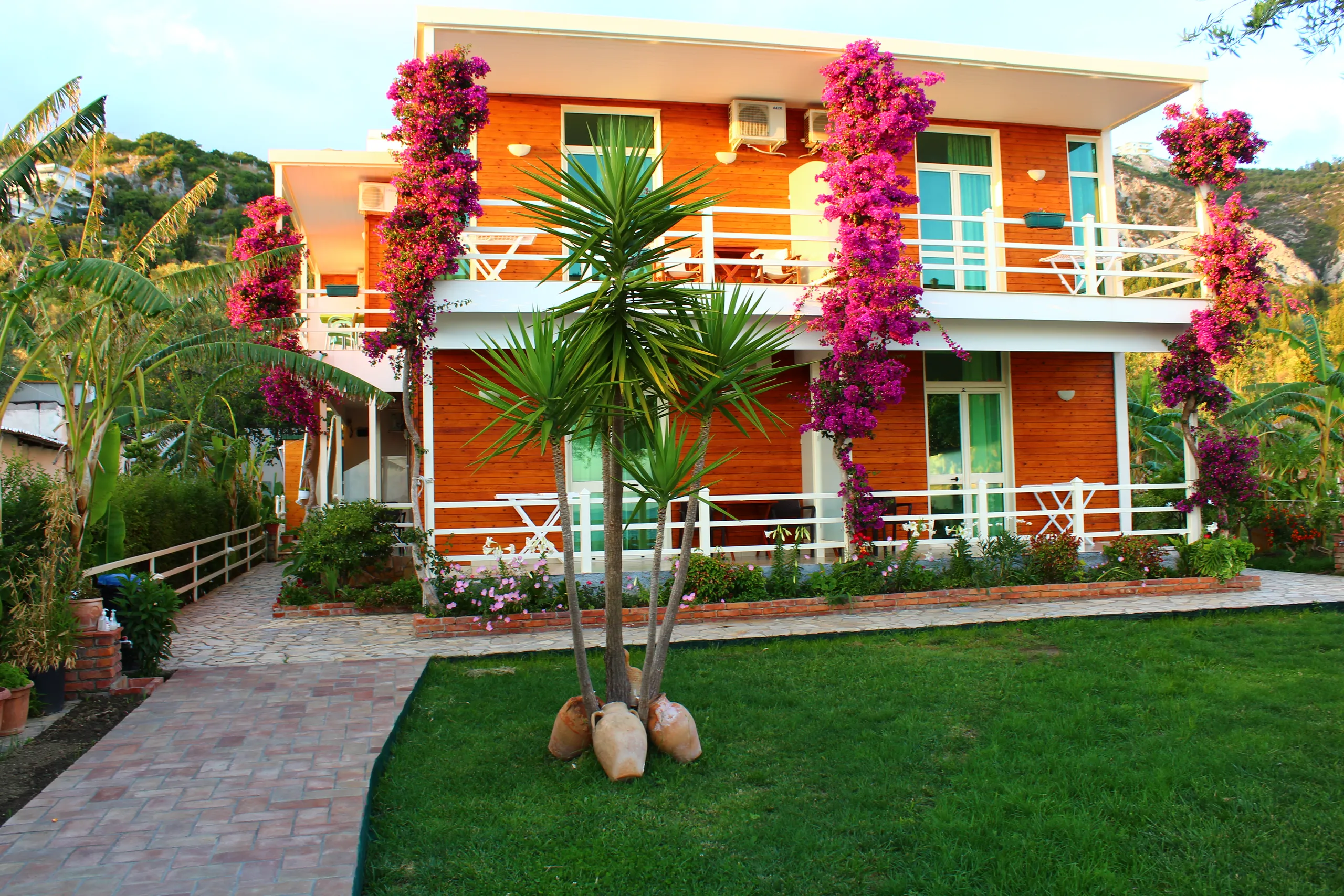 The timber house in late sun behind its lawn, bougainvillea climbing the posts and green glass doors on both floors
