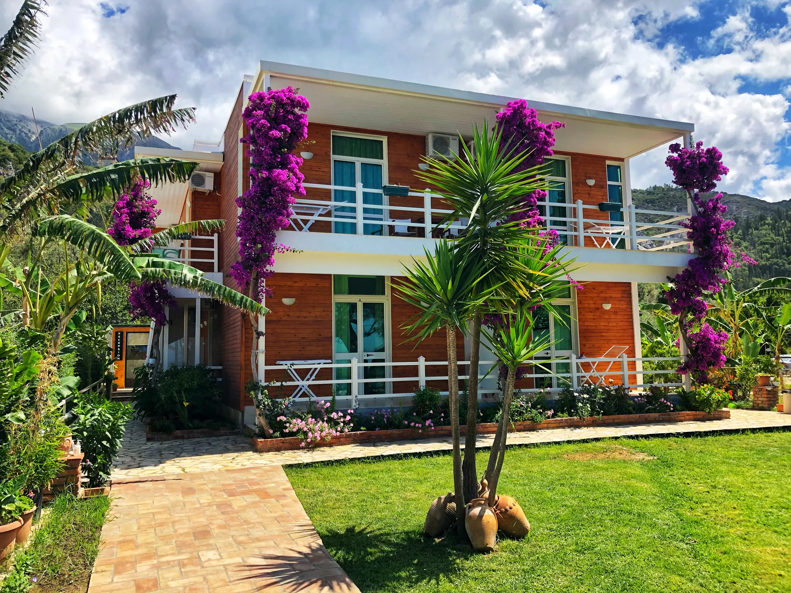 Hotel Jon: a two-storey house clad in orange-stained timber with white rails, columns of magenta bougainvillea, a yucca in clay amphorae on the lawn