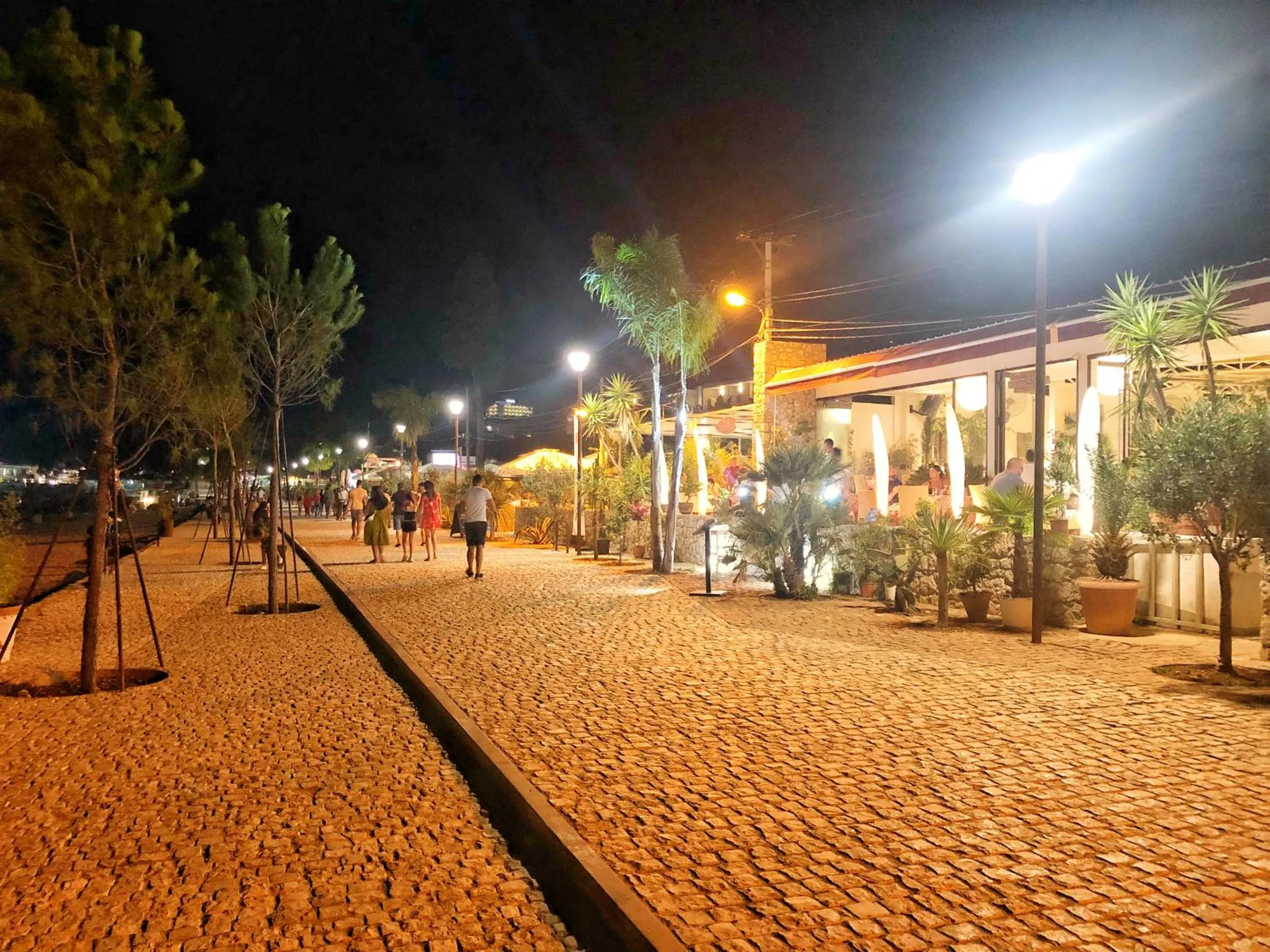 The beach promenade at night, lamps along the paving, people walking past the lit terraces