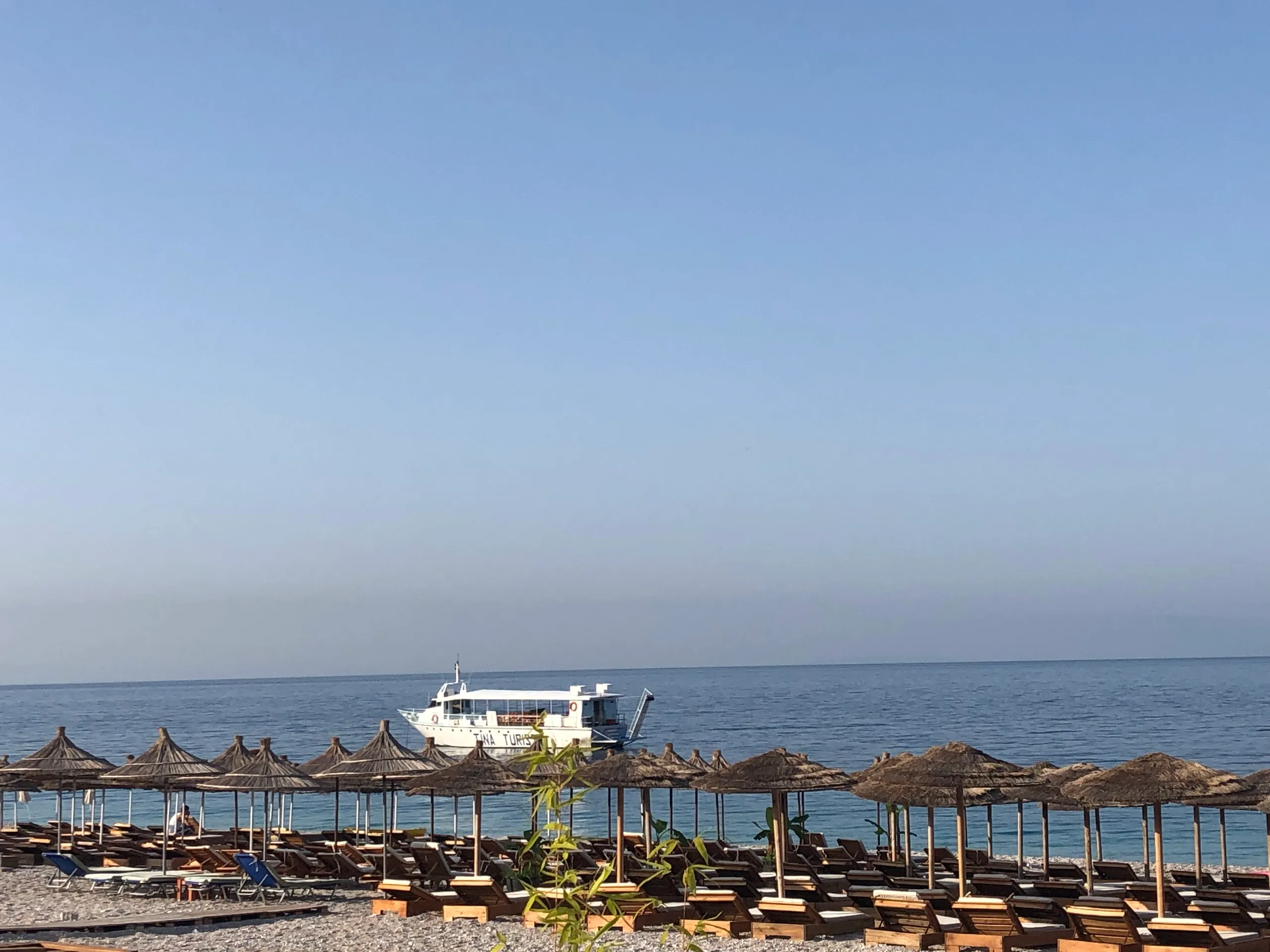 Straw umbrellas and wooden sunbeds on the pebble beach in the morning, a white boat on the calm sea