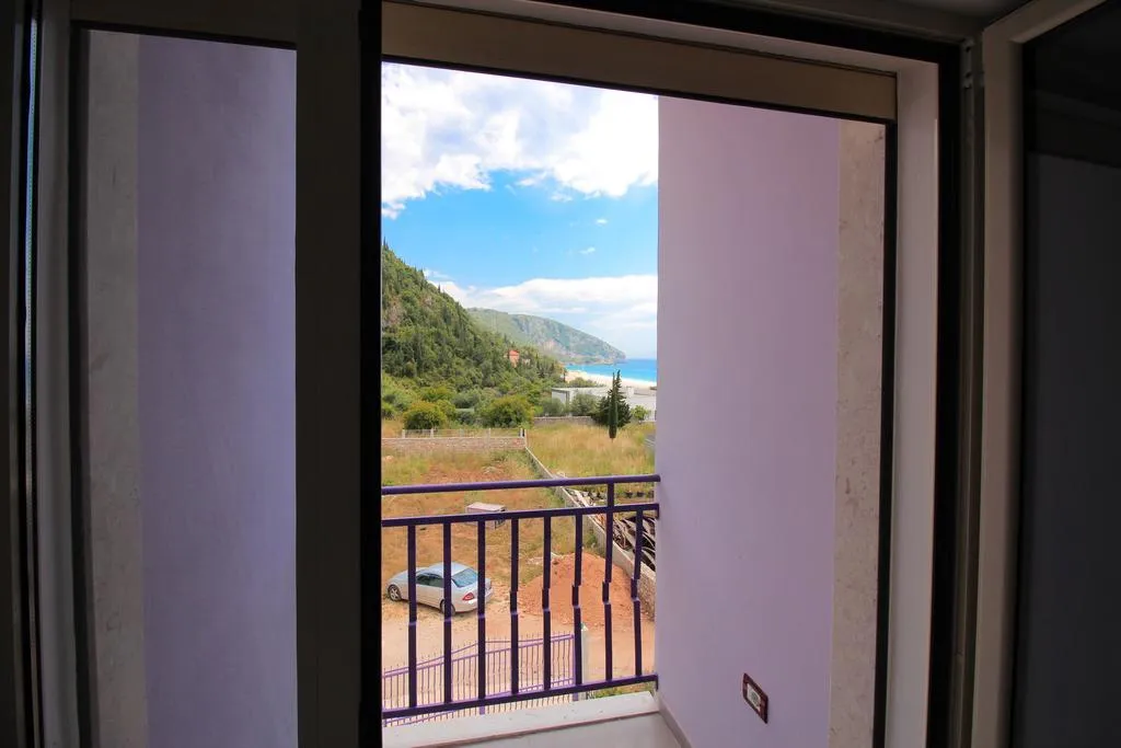 An open balcony door in a lilac-walled recess at Hotel Keisa, framing green hills, the end of the bay and a strip of sea