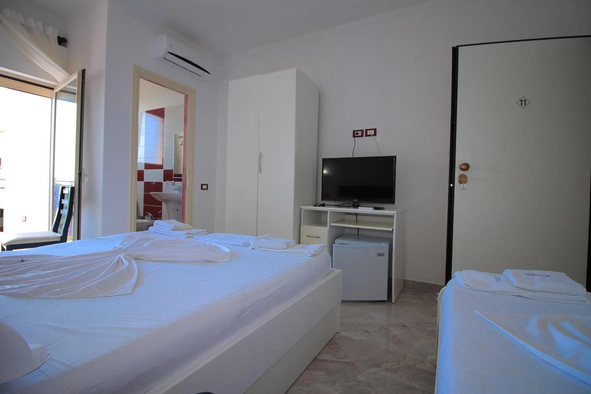 A bright room at Hotel Keisa with a double bed and a single bed, a white wardrobe, a TV on a white stand, a small fridge and an air conditioner over the door