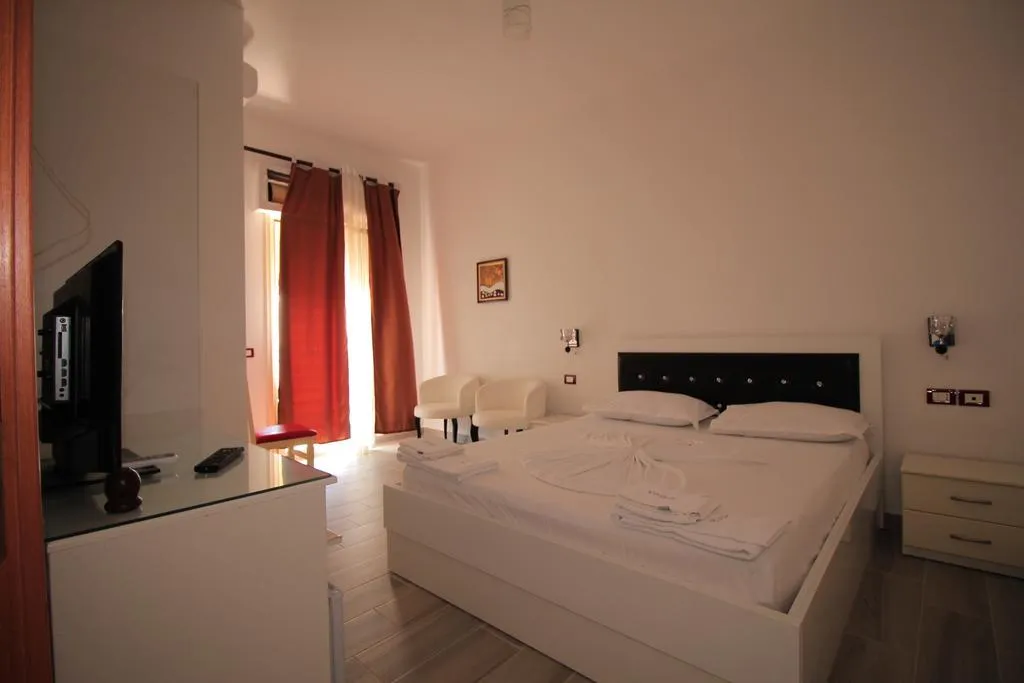 A double room at Hotel Keisa with a white bed and black quilted headboard, two white armchairs, red curtains at the balcony door and a flat-screen TV