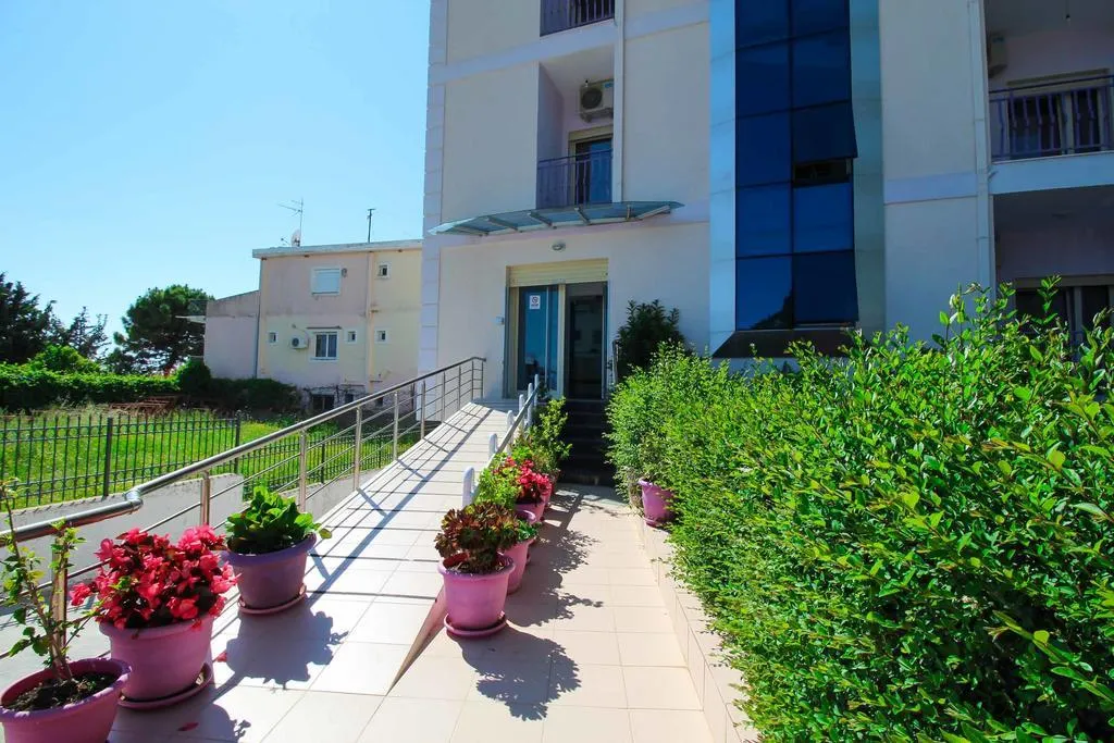 The entrance path to Hotel Keisa lined with pink flower pots and a white rail, leading to the door under the blue glass column