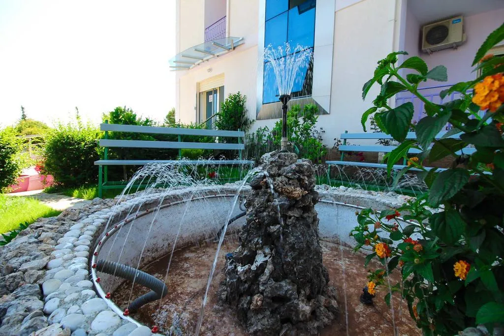 A round stone fountain on a lawn in the front garden of Hotel Keisa, with flowering shrubs, benches and the cream building with its blue glass column behind