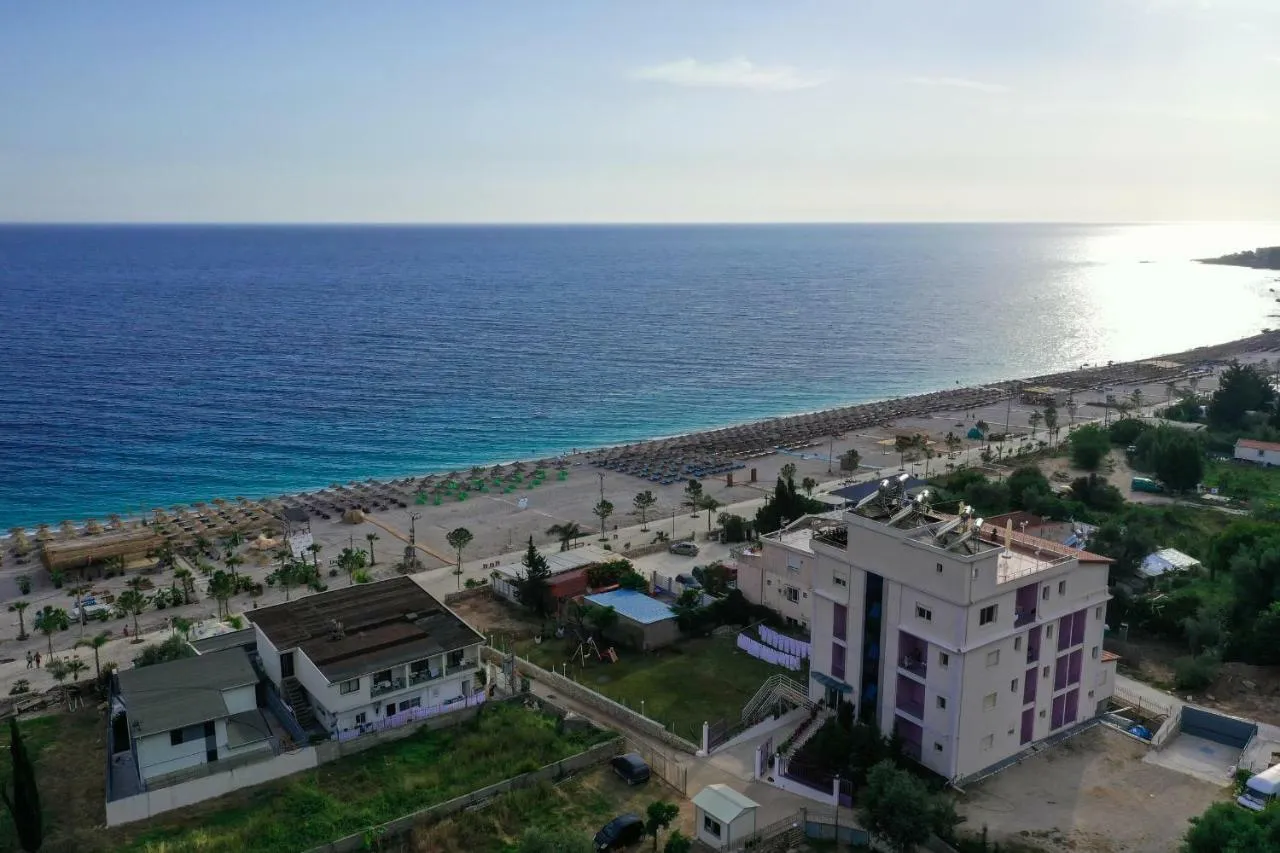 An aerial view over the sea and Dhërmi beach: rows of umbrellas, the promenade with palms, and behind them the tall cream-and-lilac block of Hotel Keisa with its blue glass column