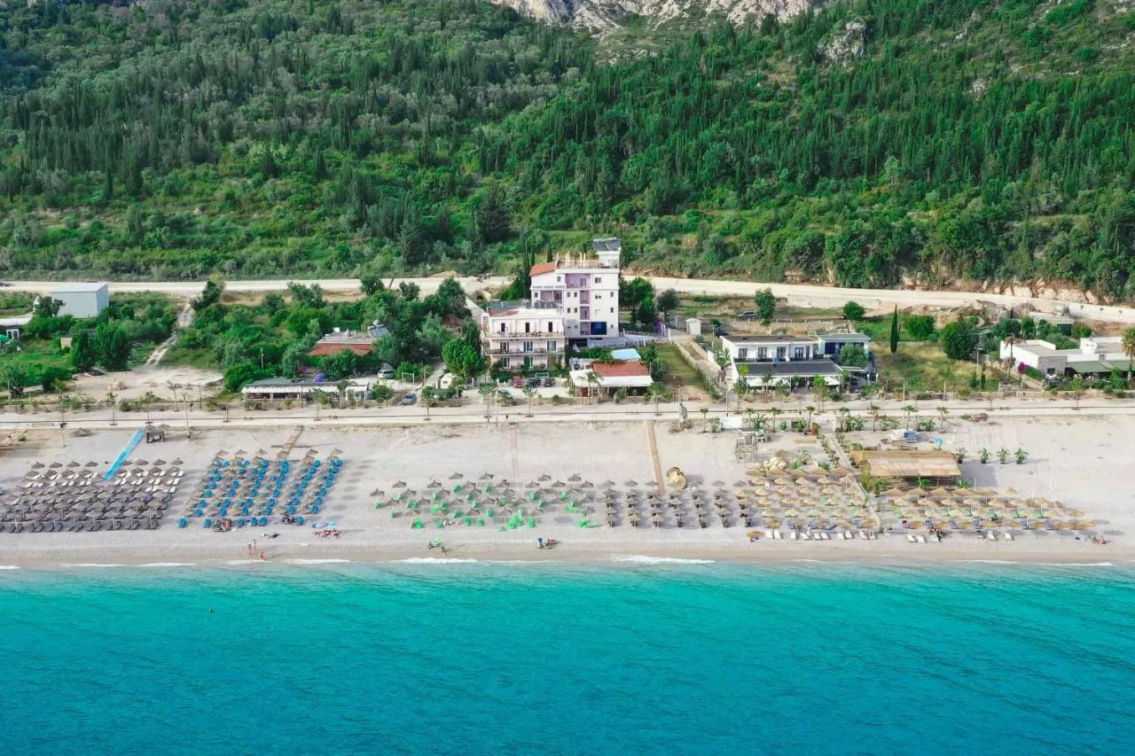 An aerial view of turquoise water, the umbrella rows of Dhërmi beach and the tall pale building of Hotel Keisa among low roofs, with pine-covered slopes rising behind