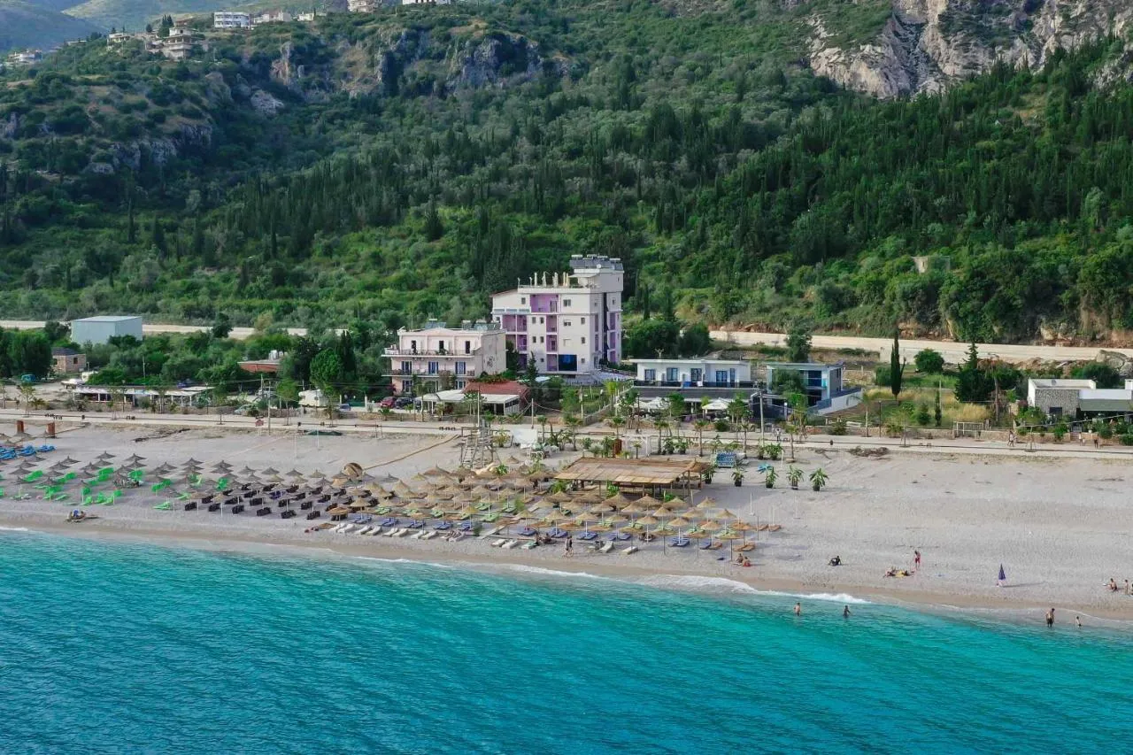 An aerial view from over the water of Dhërmi beach with its umbrellas and the tall cream building of Hotel Keisa behind, under a green mountainside