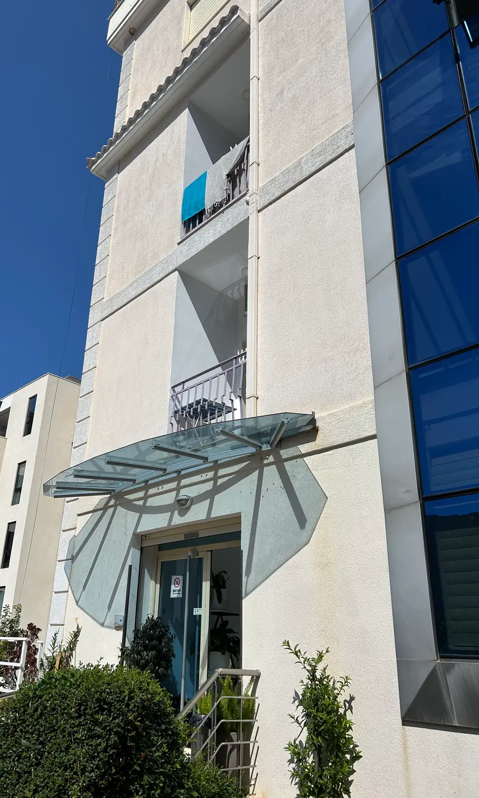 The front of Hotel Keisa looking up: cream render, a frosted glass canopy fanned over the entrance door, white balcony rails above it and a full-height column of cobalt glass panes on the right against a clear blue sky