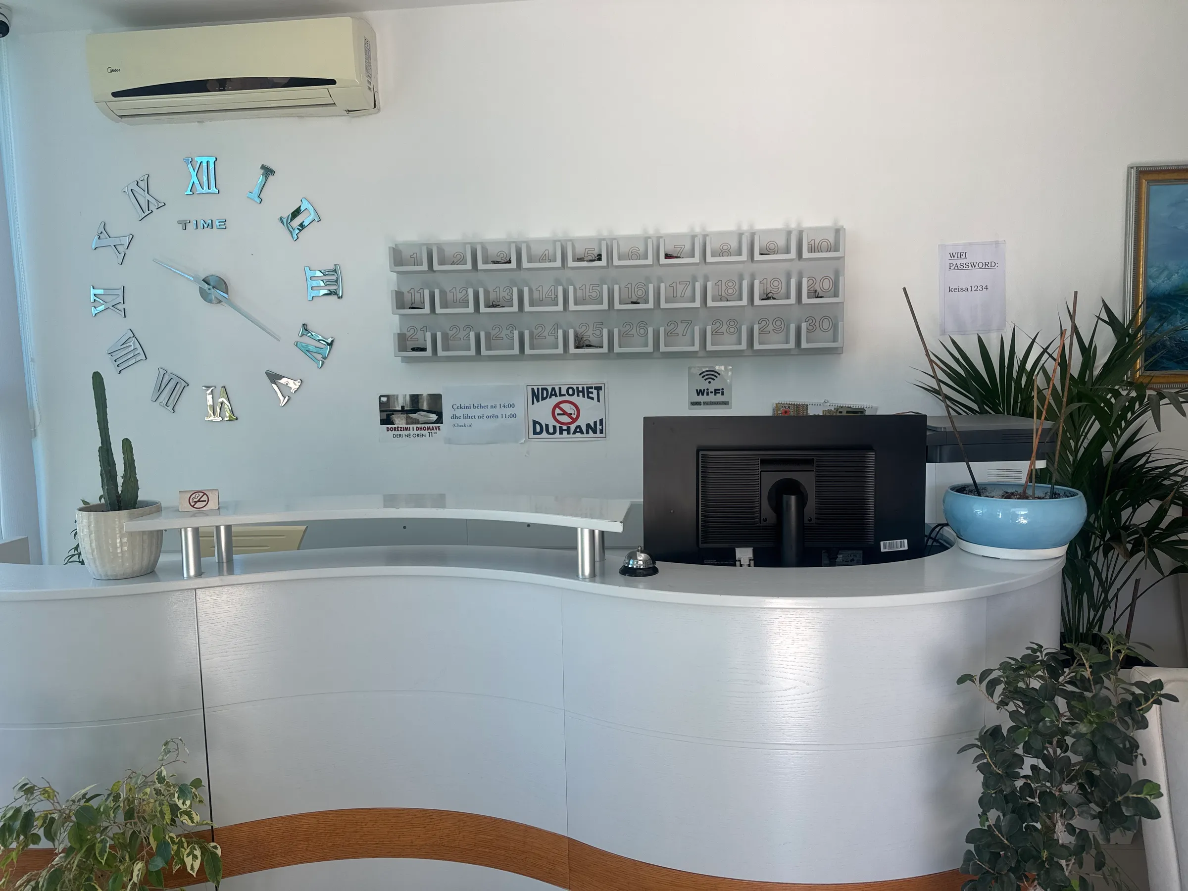The curved white reception desk at Hotel Keisa with a Roman-numeral wall clock, a rack of room-key boxes and potted plants