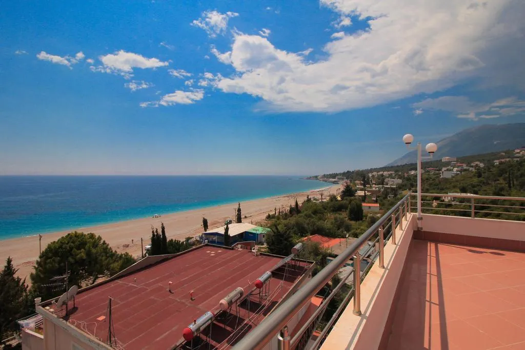 The terracotta-tiled roof terrace of Hotel Keisa behind a steel rail with globe lamps, looking over rooftops to the long pale curve of Dhërmi beach and the sea