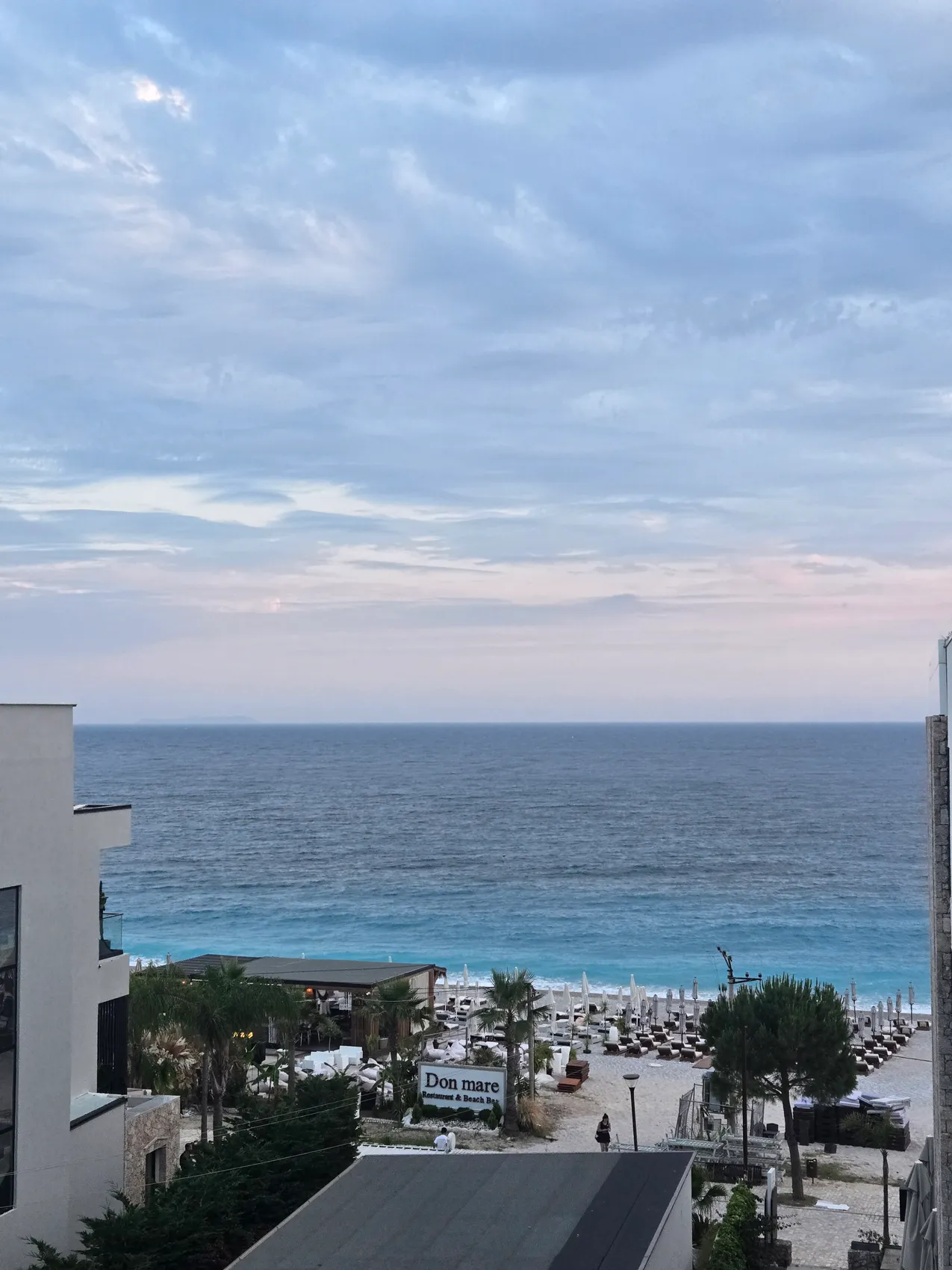 A view from an upper floor down between buildings and pine trees to the beach bars and umbrellas of Dhërmi beach and the open sea under a pale evening sky