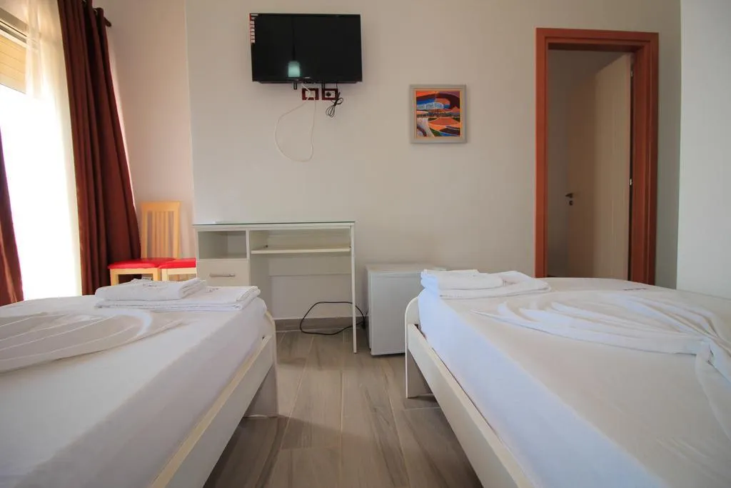 A twin room at Hotel Keisa with two white single beds, a wall-mounted TV, a white desk and red curtains at the window