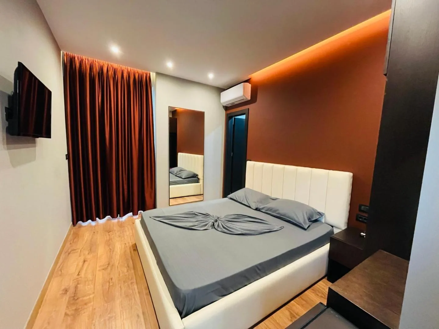 A room with grey bedding on a white padded headboard, a rust-red floor-length curtain, a wall-mounted television and a mirror