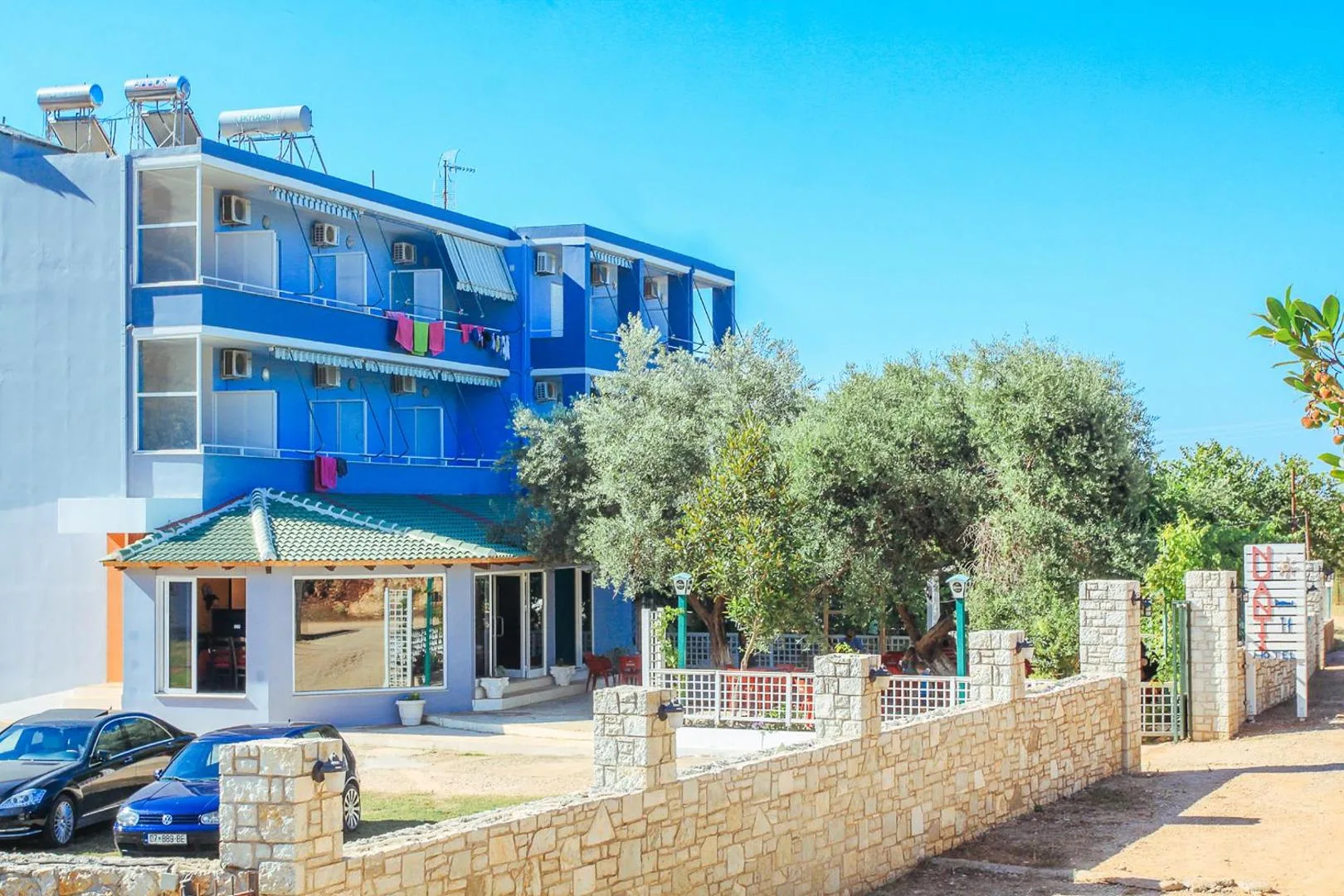 A three-storey hotel painted blue and white, standing among olive trees above a walled parking court