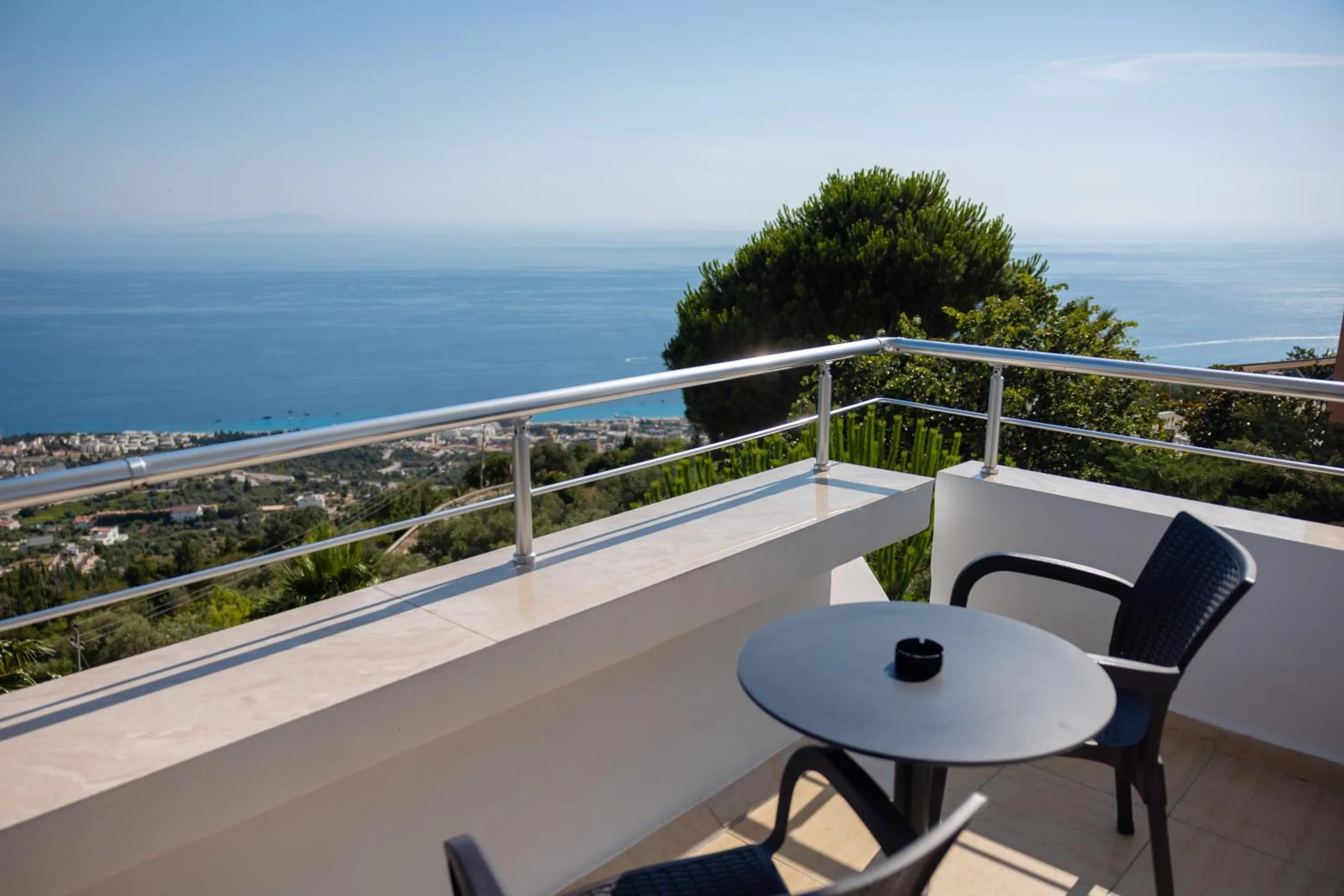 A round table and two chairs on a balcony with a stainless rail, a stone pine and the bay below