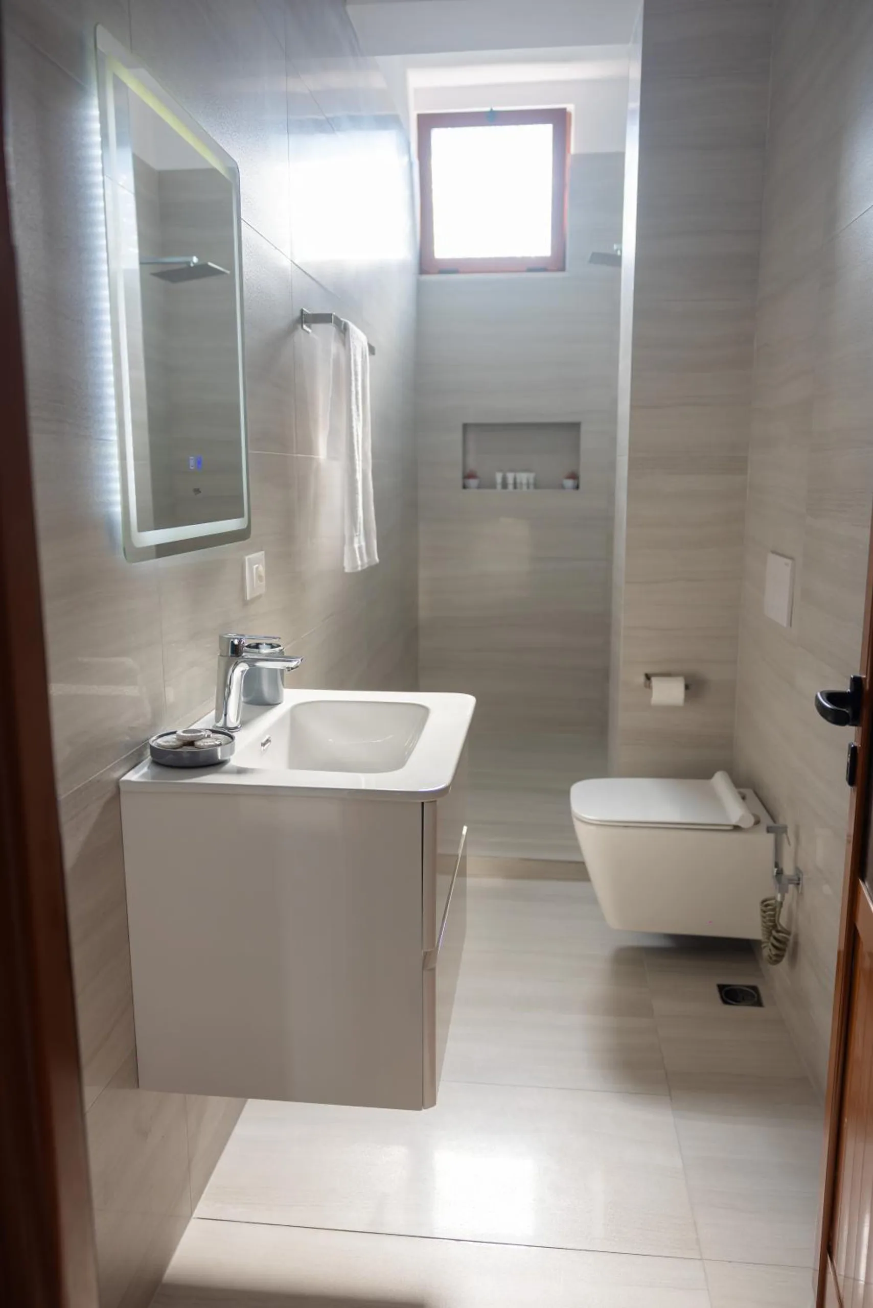 A shower room in pale grey stone-effect tile with a wall-hung basin, a lit mirror and a small window