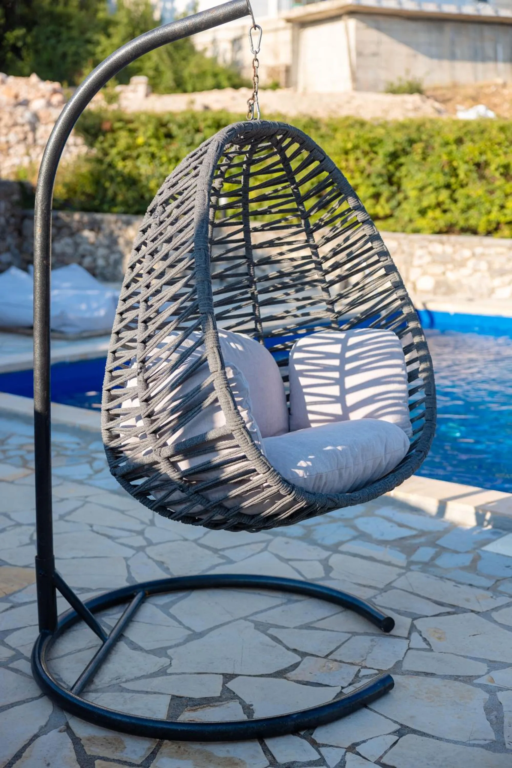 A grey wicker hanging chair with cushions on the paved poolside