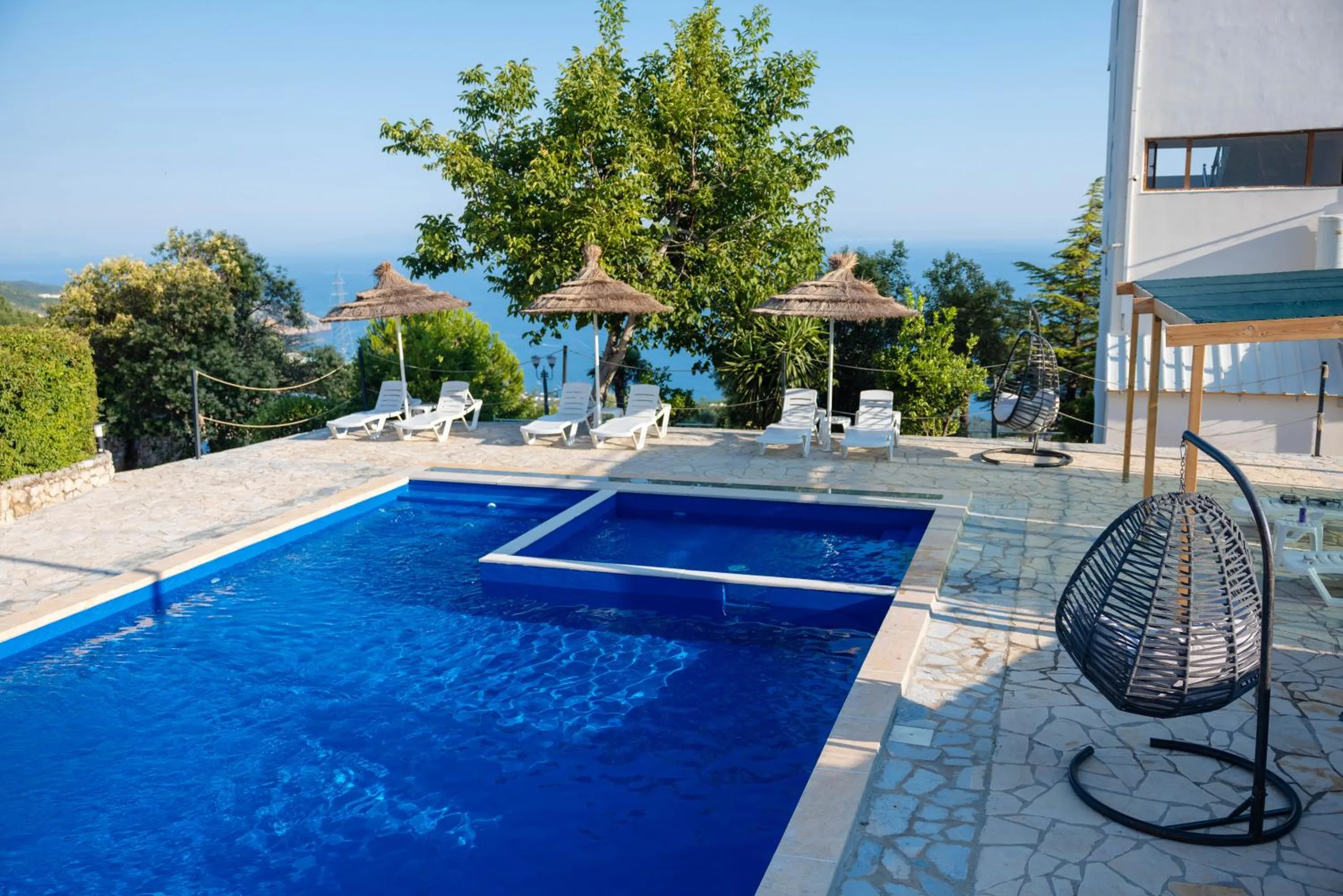 The pool beside the white hotel wall, a hanging wicker chair and parasols with the sea showing through the trees