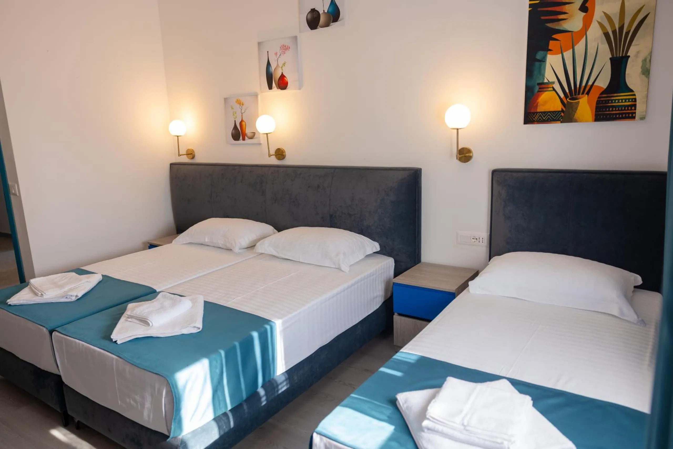 A room with two beds side by side, slate velvet headboards, globe wall lamps and bright paintings