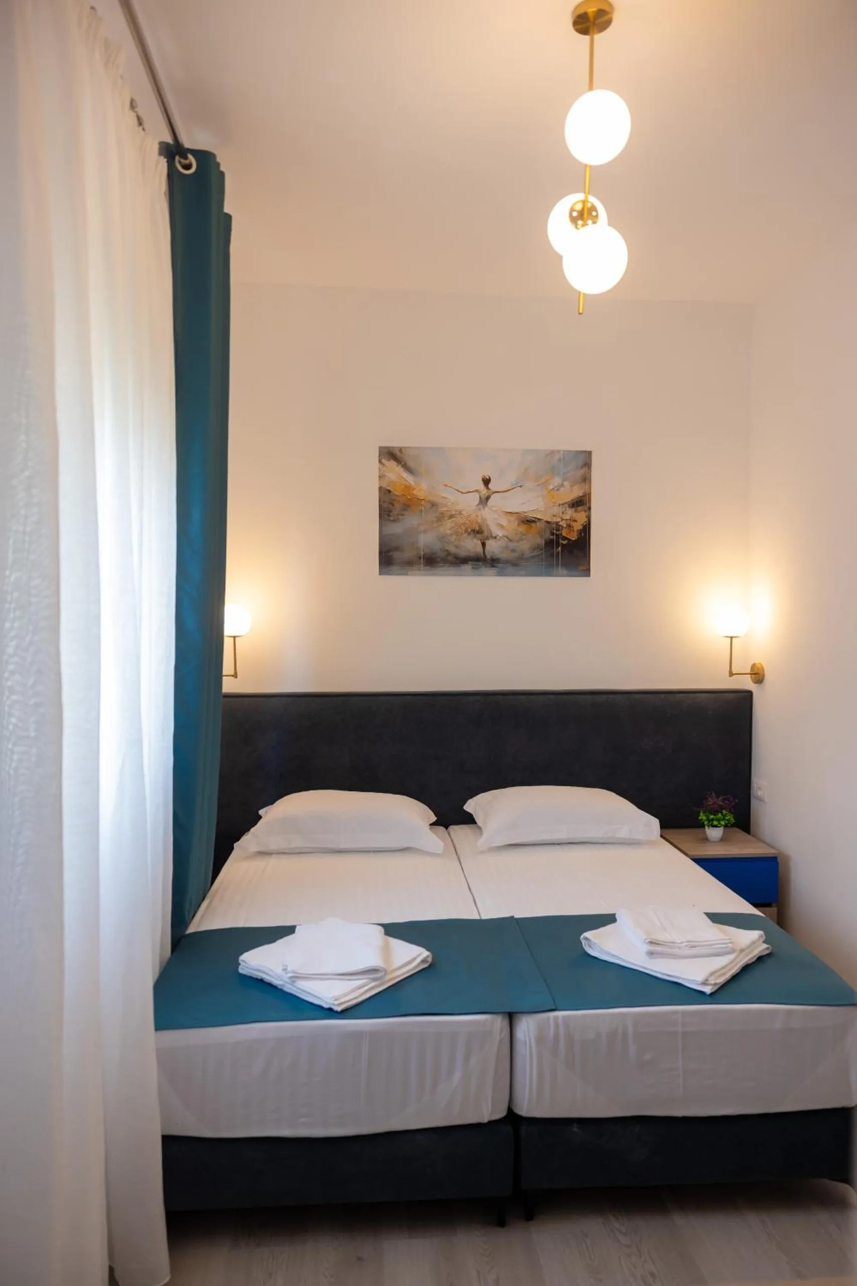 A narrow room with a double bed, a pendant of glass globes and a painting of a dancer above the bed