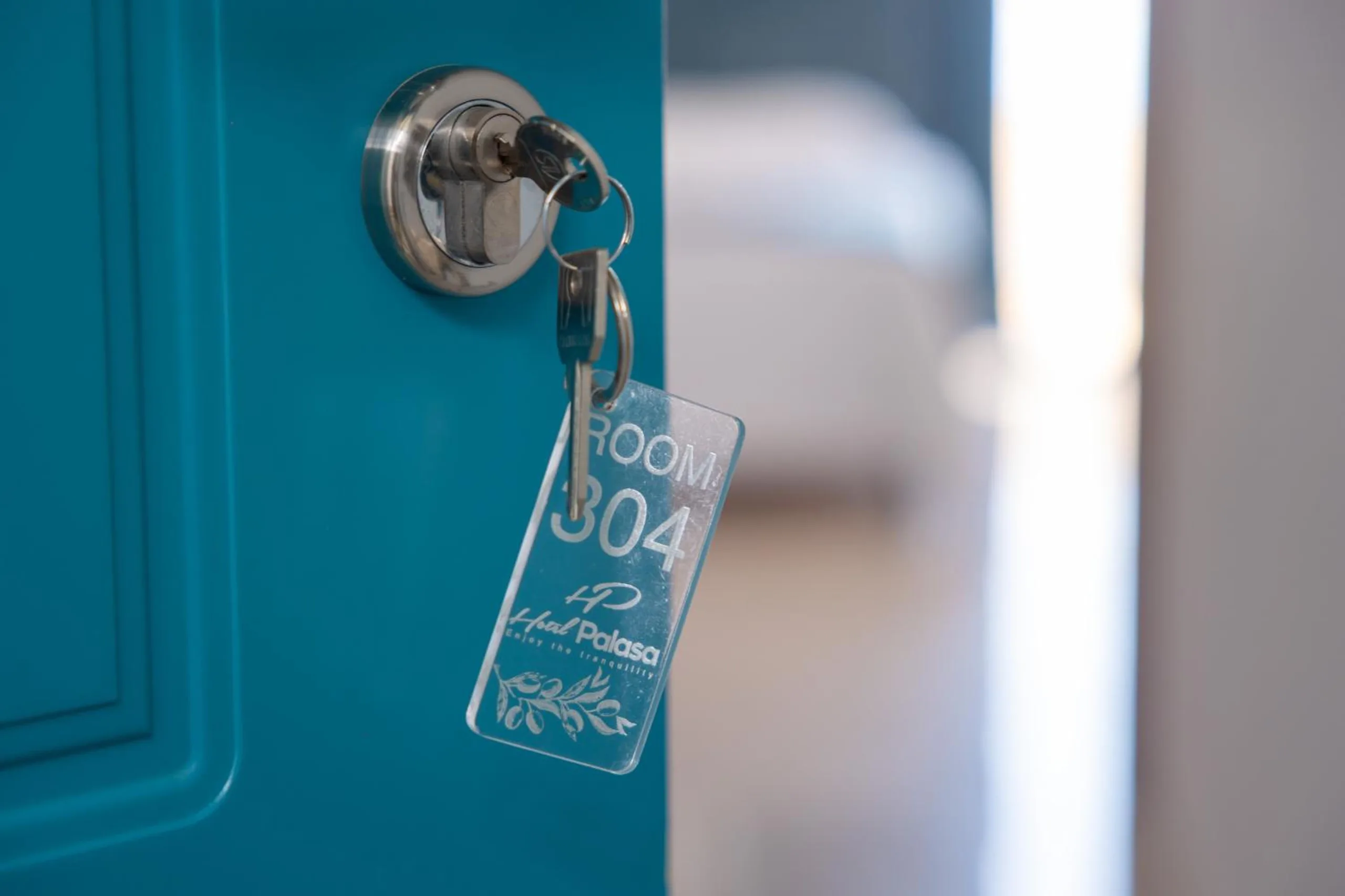 A key in the lock of a turquoise door on a clear tag engraved Room 304, Hotel Palasa, with an olive sprig