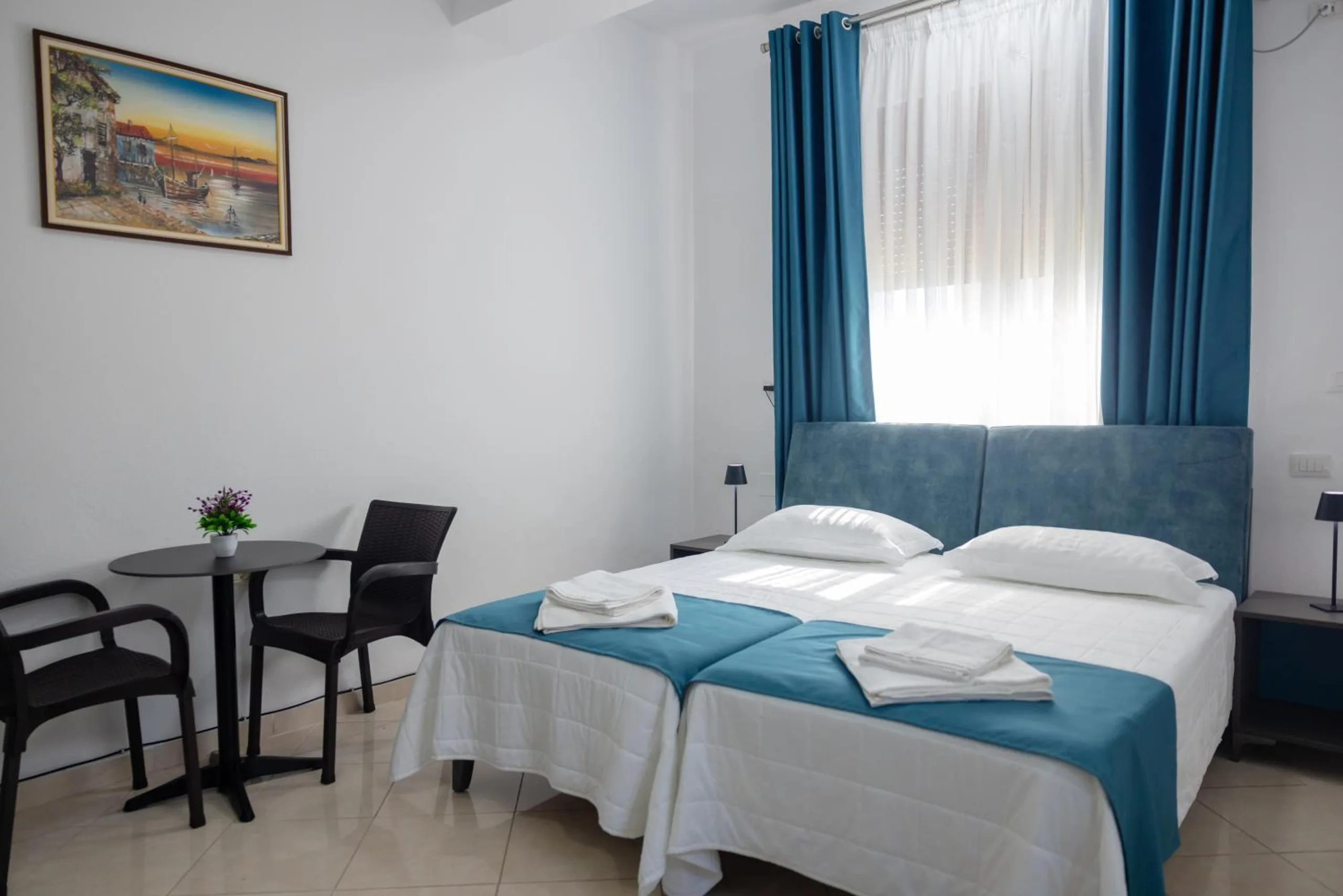 A bright room with two beds joined, teal runners, a small round table with two chairs and long teal curtains