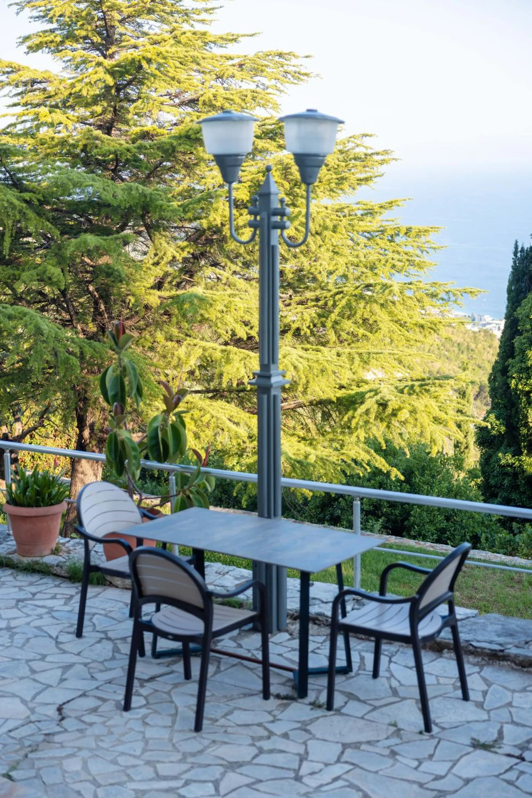A table for four on a stone terrace beside a lamp post, with a cedar and cypresses and the sea behind