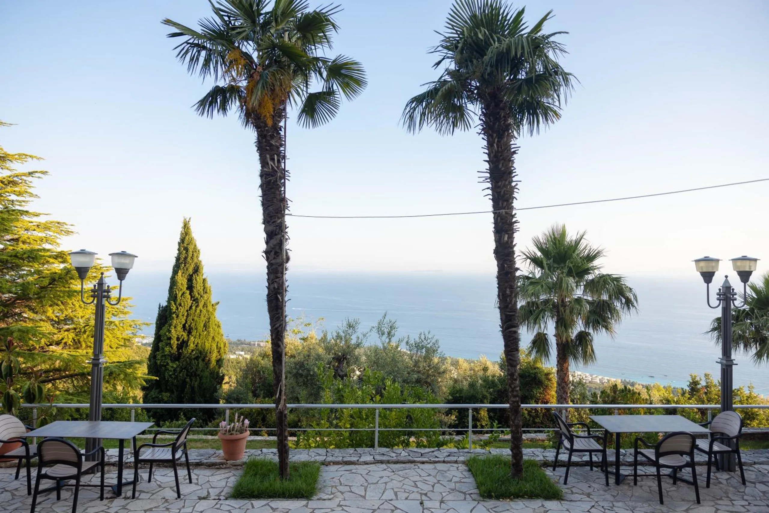 Tables and chairs on a crazy-paved terrace between two tall palms, street lamps and the sea beyond