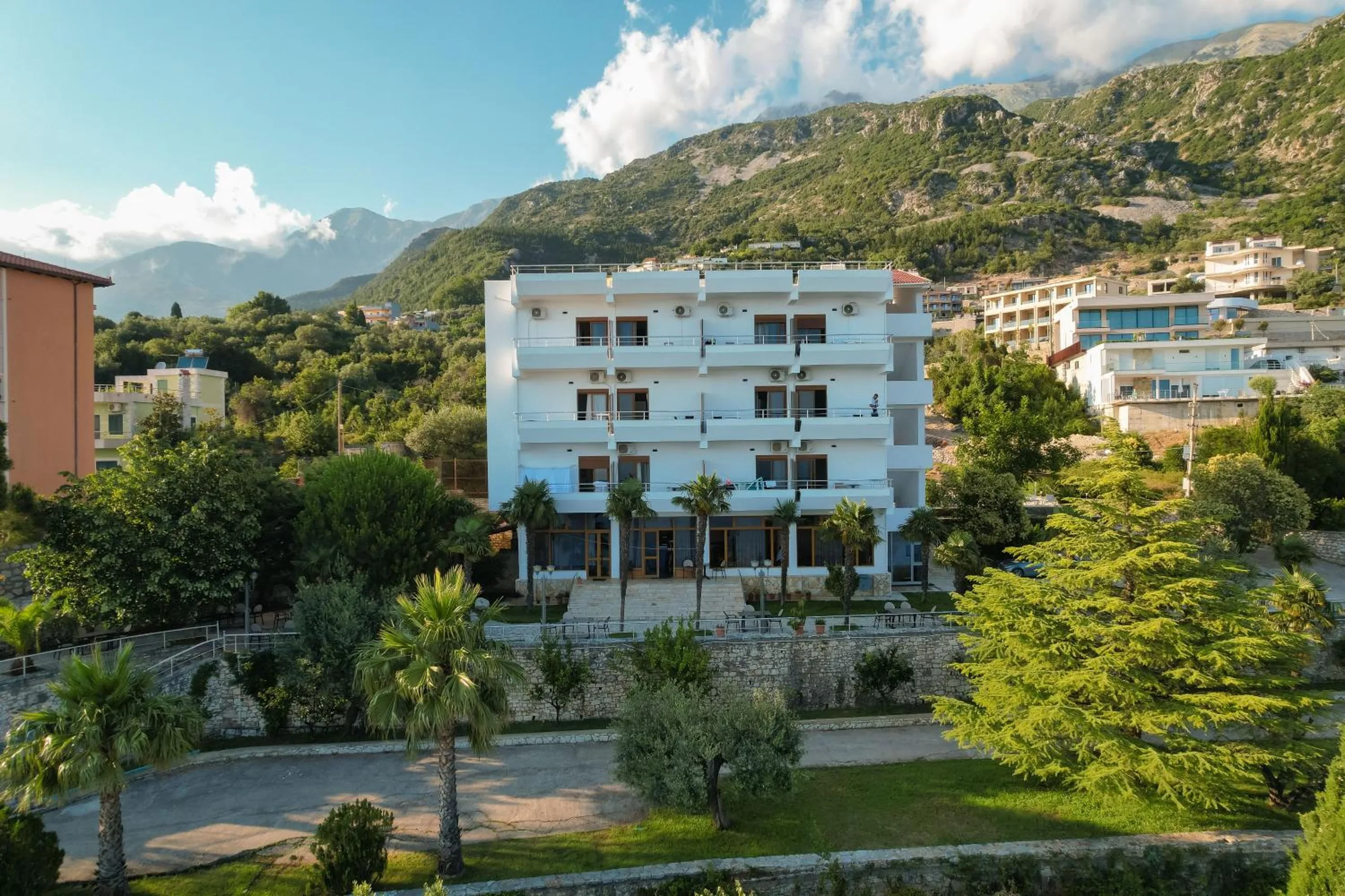 The white four-storey hotel building with balconies, palms and a lawn in front and mountains behind