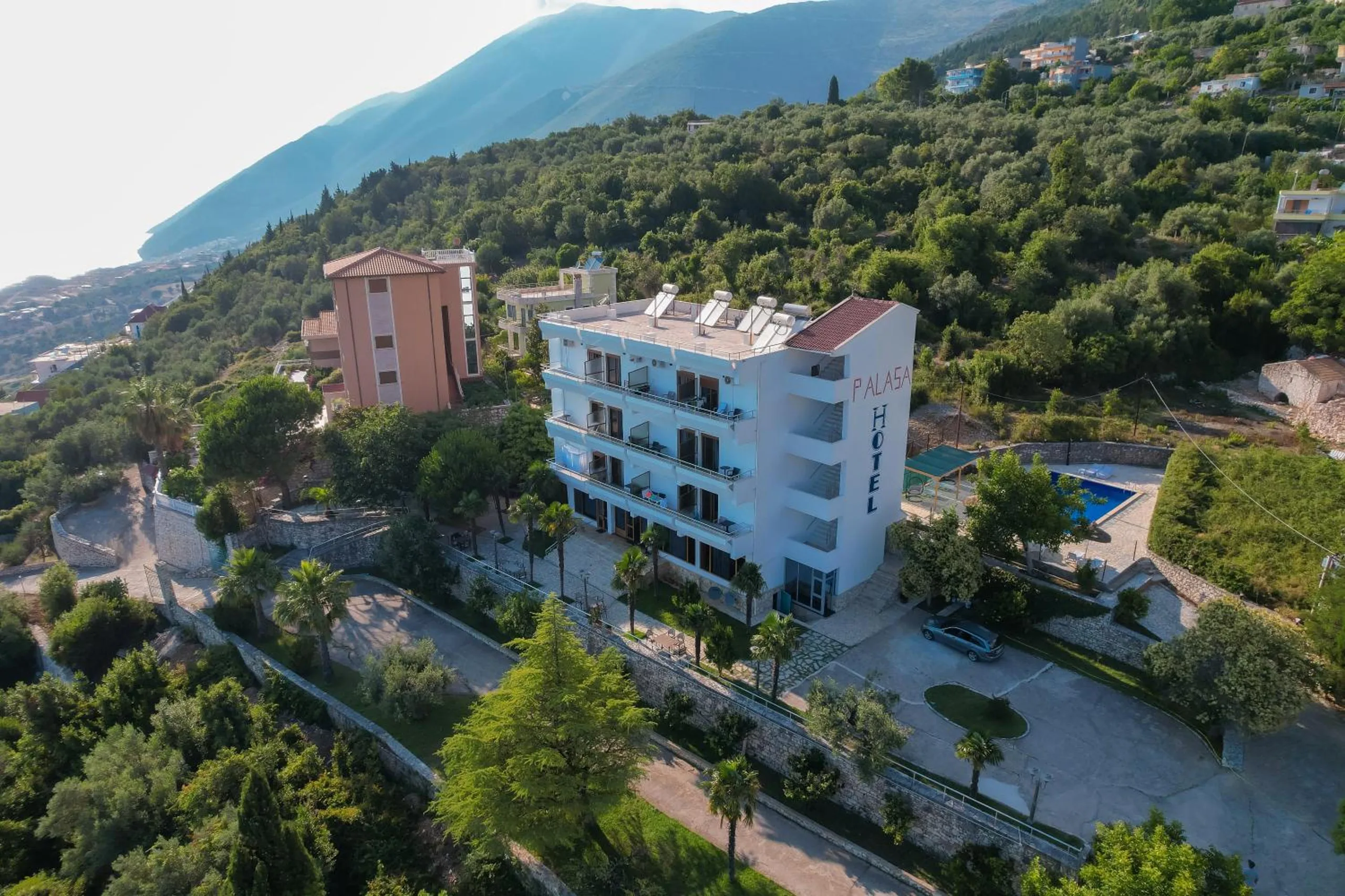 An aerial view of Hotel Palasa with its painted name on the gable, the pool beside it and the wooded hillside rising behind