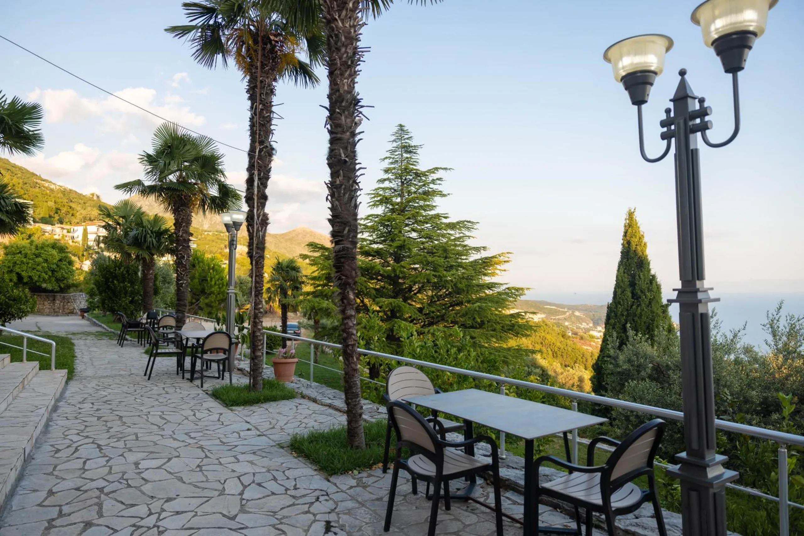 A paved terrace path lined with palms, tables and a double street lamp, the coast below