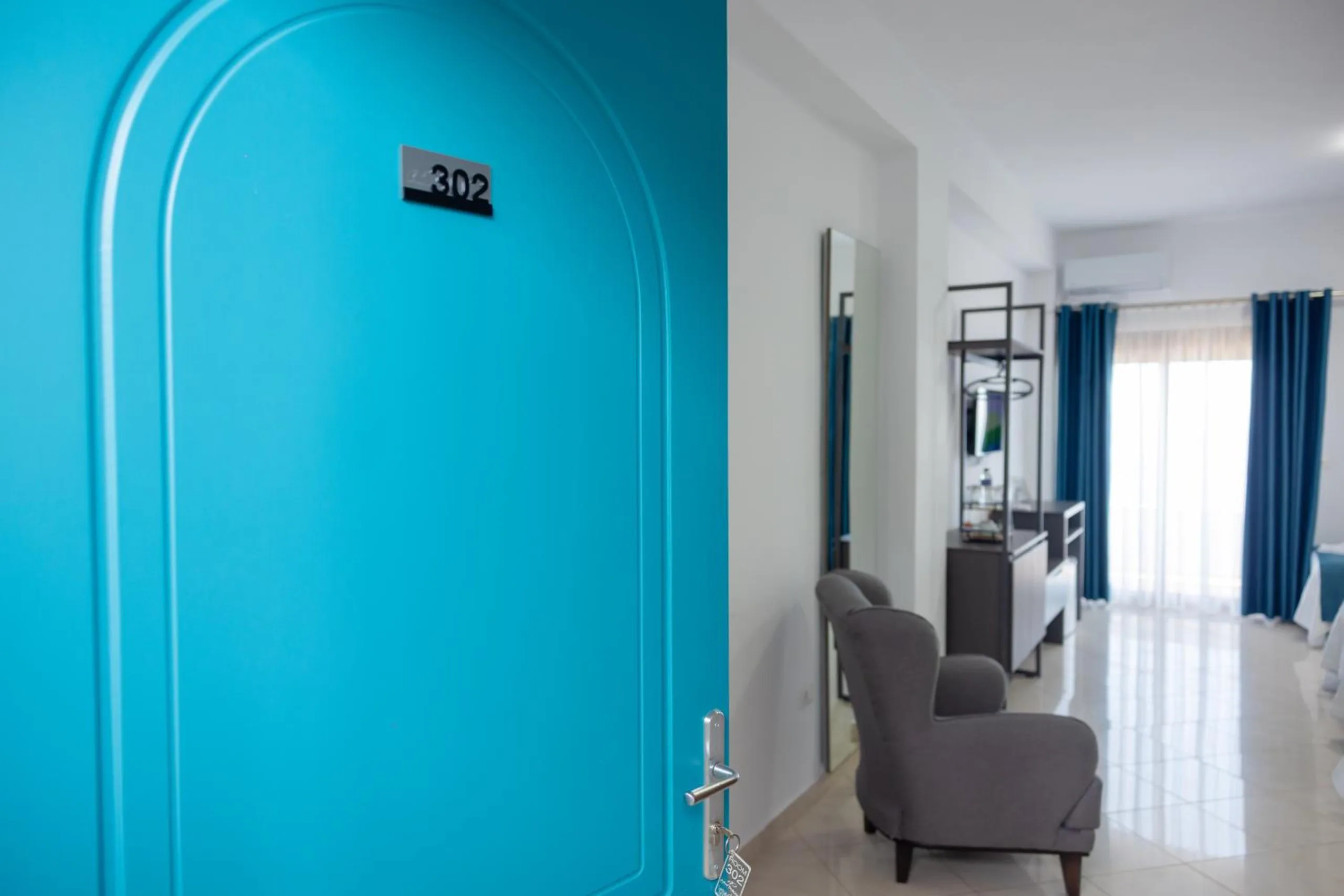 A turquoise panelled door numbered 302 open onto a room with a grey armchair and blue curtains