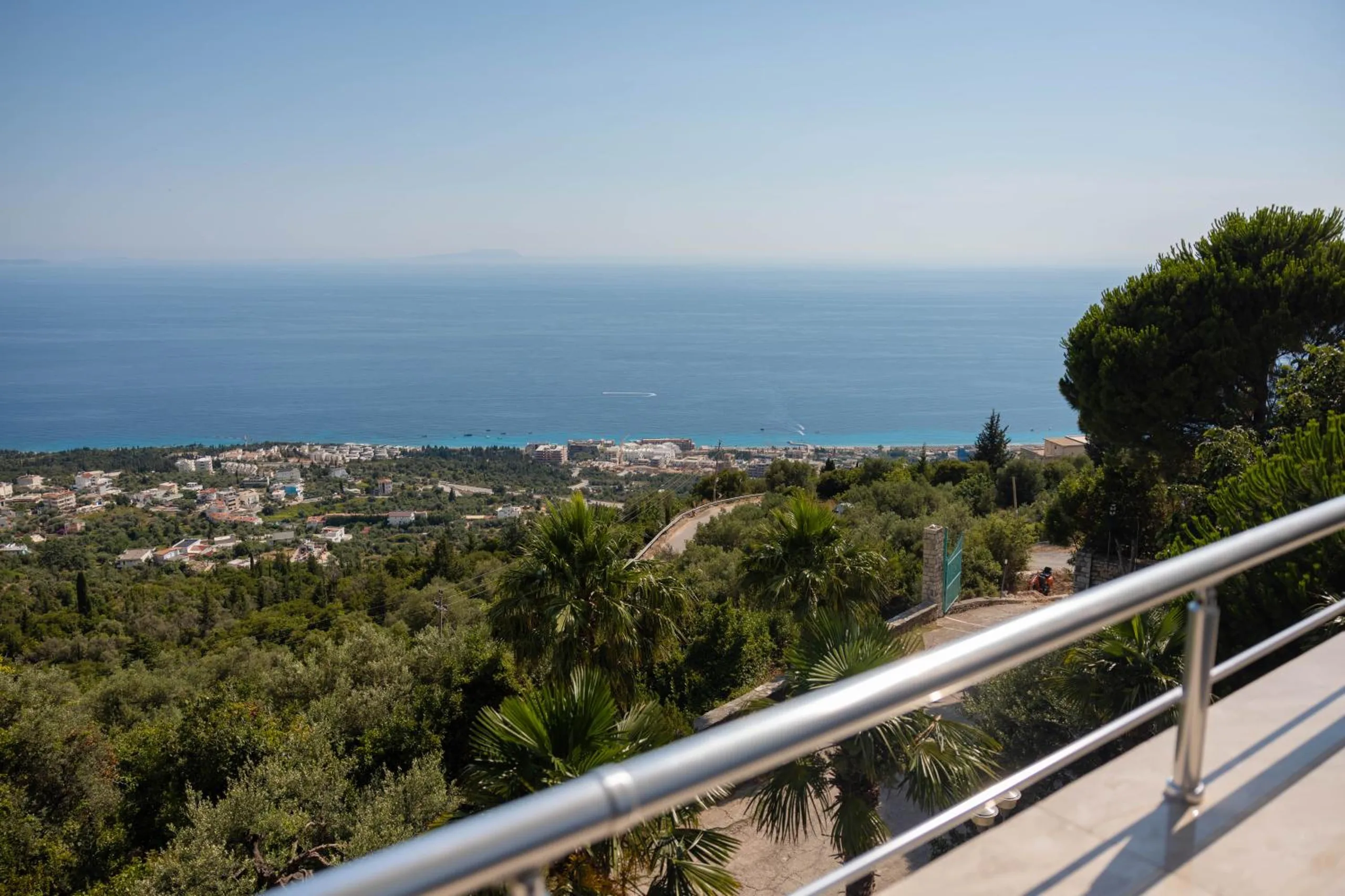 The view from a balcony rail at Hotel Palasa over olive and palm trees down to Drymades and the Ionian Sea, an island faint on the horizon