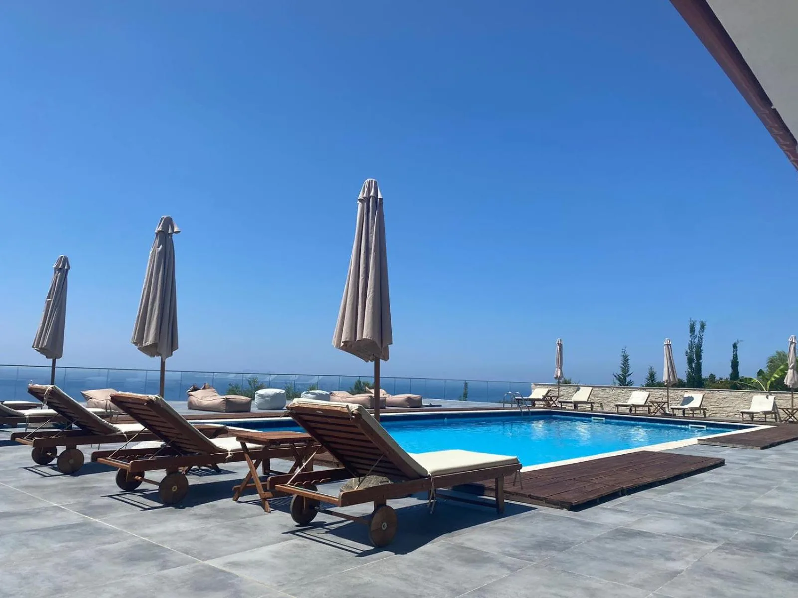 The pool at Hotel Relax Dhermi under a clear blue sky, wooden loungers and closed umbrellas on grey stone tiles, the sea beyond a glass balustrade