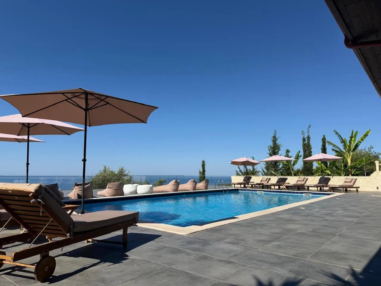 Open sand-coloured umbrellas and loungers around the pool, bean bags along the balustrade and the sea on the horizon