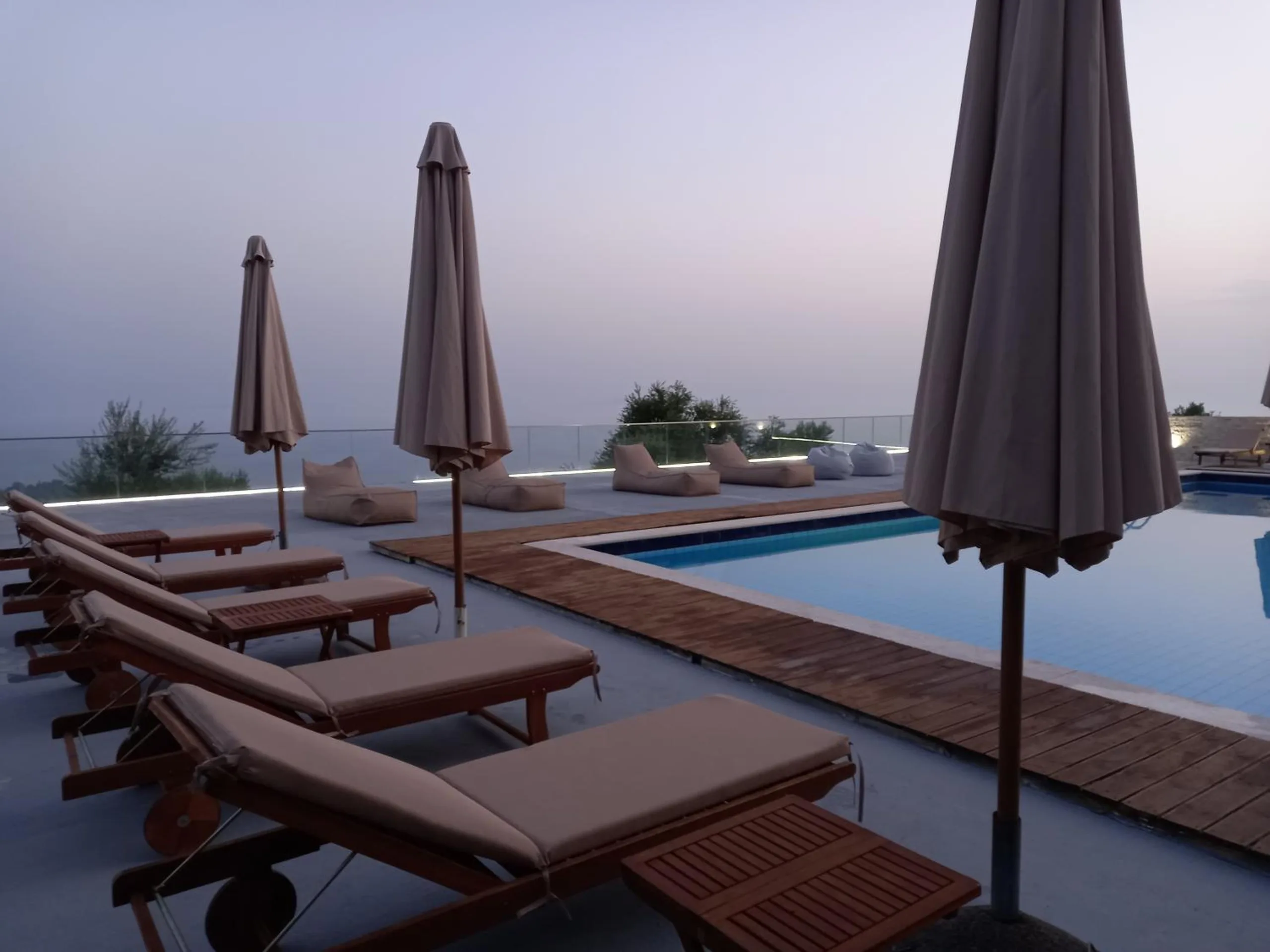 A row of wooden loungers and furled umbrellas beside the pool at dusk, the sea and sky the same lavender grey