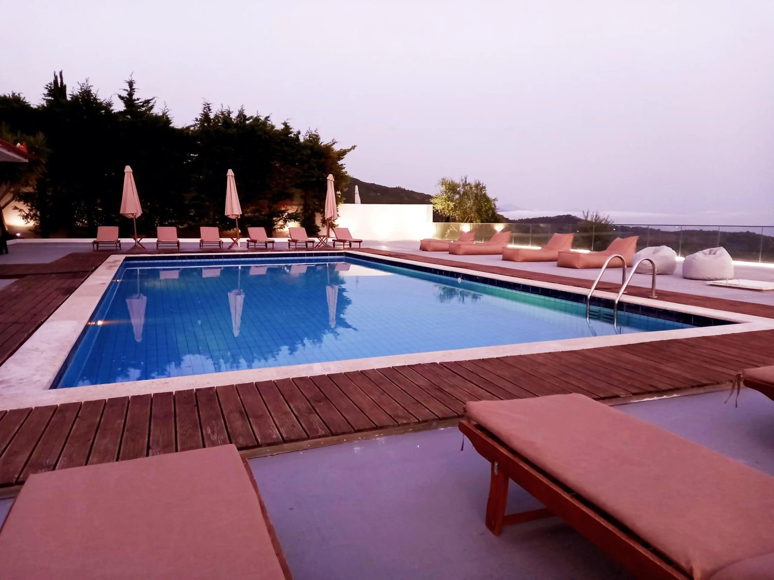 The pool at dusk framed by timber decking, umbrellas reflected in the water and pine trees behind
