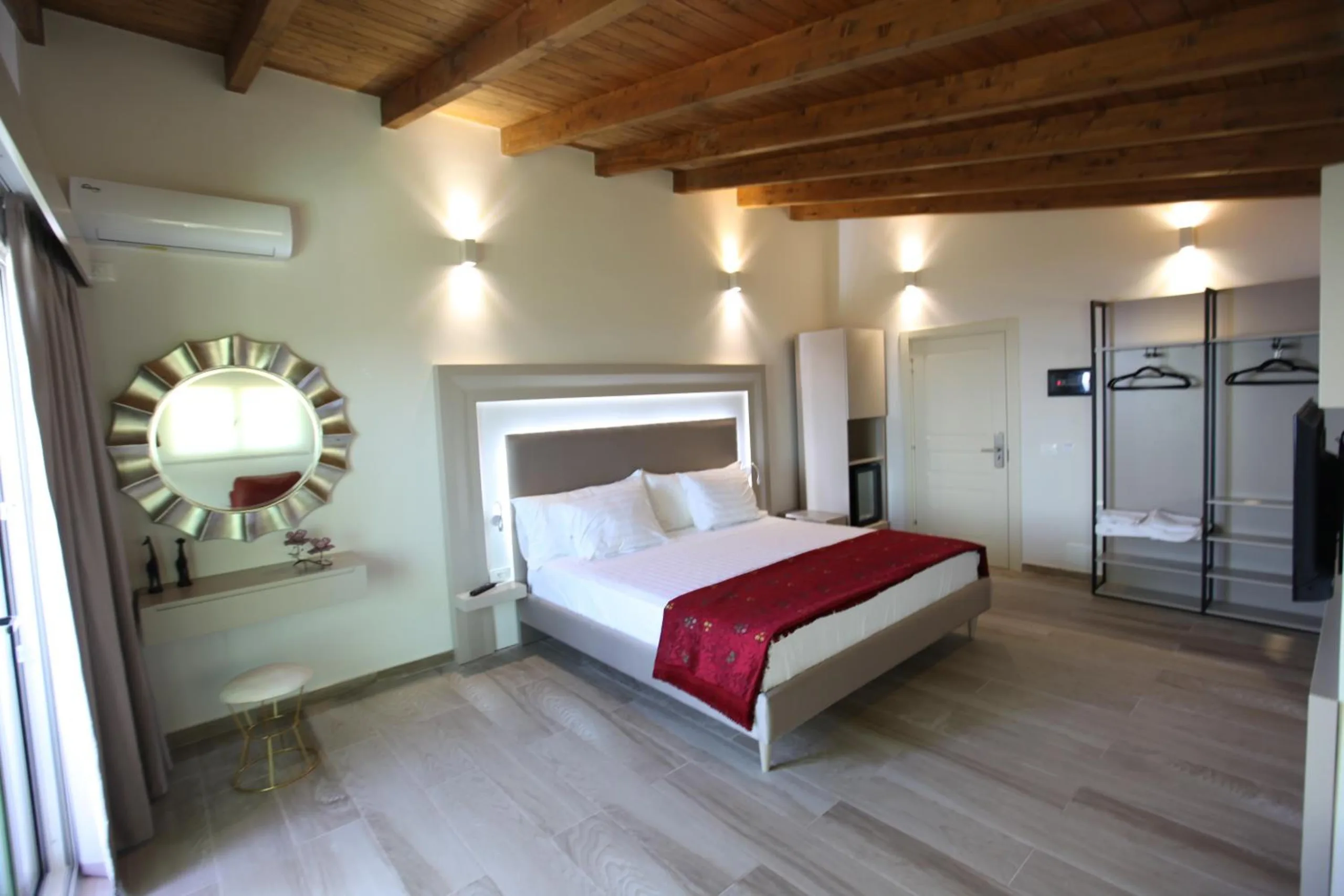 A double bed with a lit grey headboard and a red runner under a pine ceiling with exposed beams, a sunburst mirror on the wall