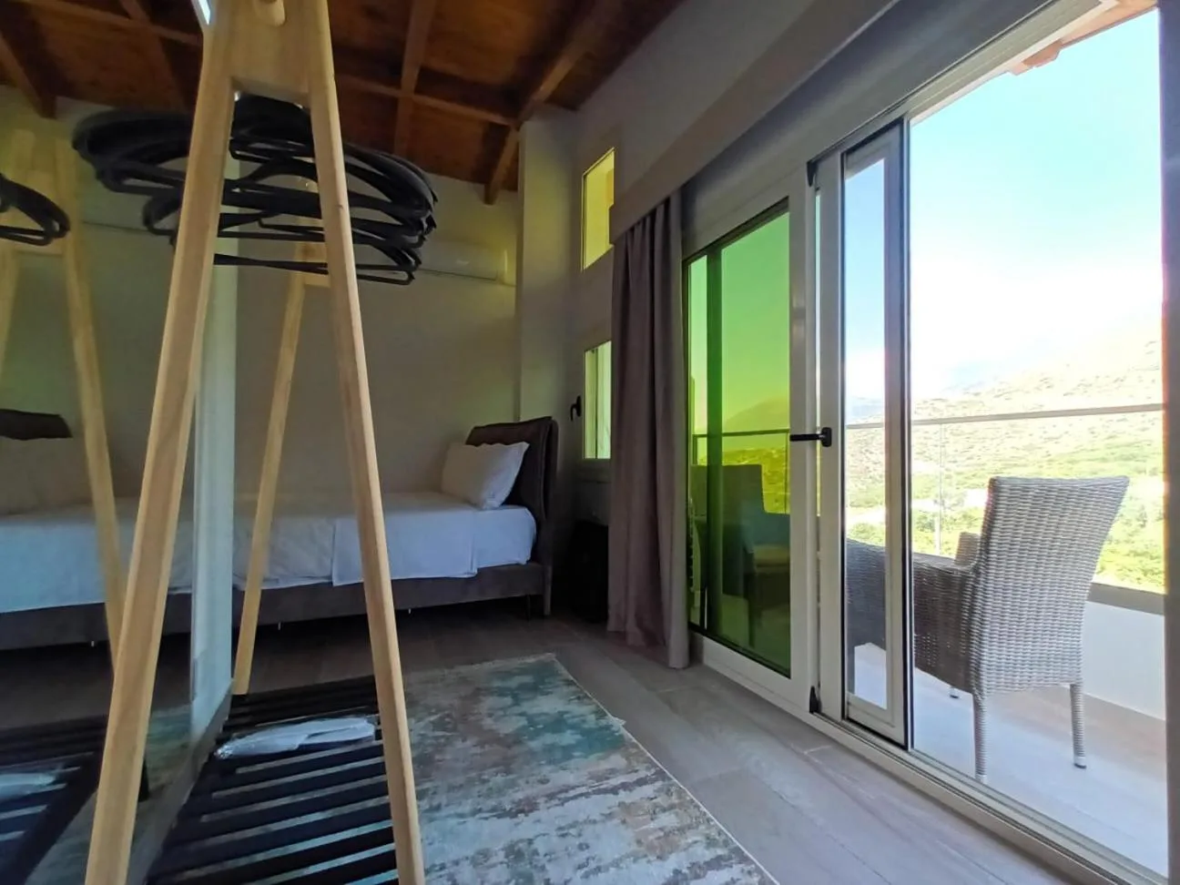 An attic room with a pine ceiling, a wooden hanger stand and a glass door to a balcony over the green hills