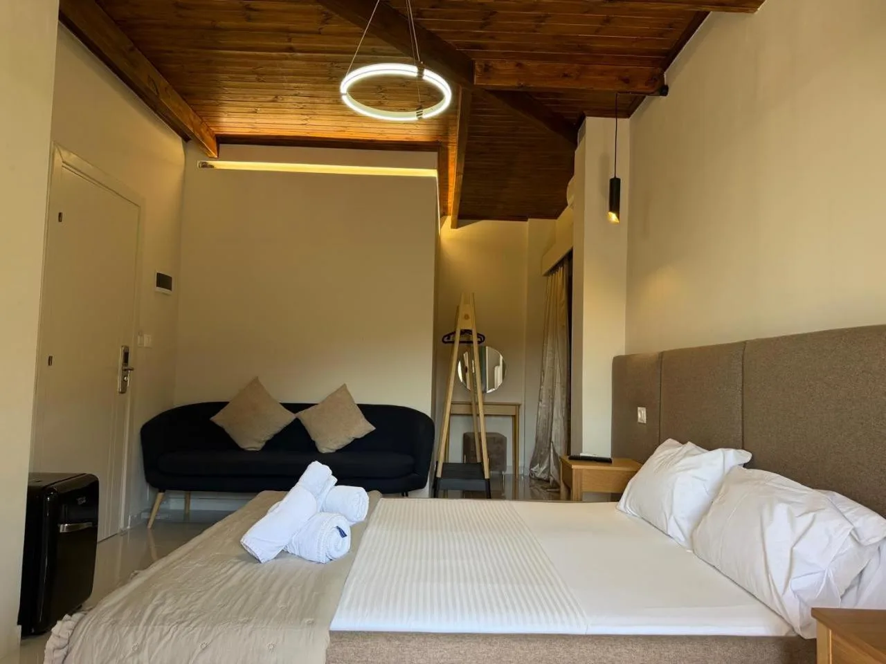 A double bed with white towels folded on it, a black sofa and a ring-shaped ceiling light under timber beams