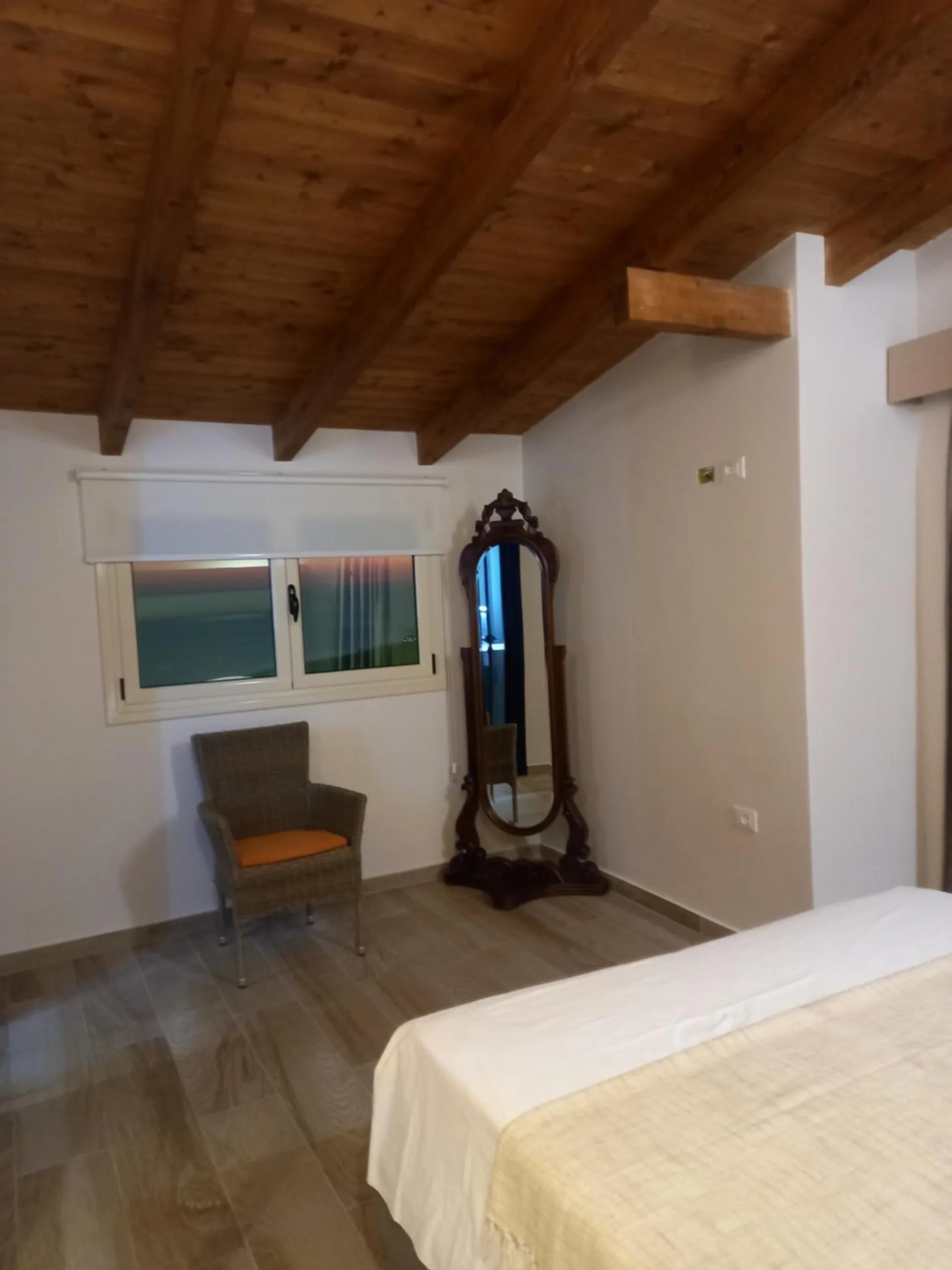 An attic room with a pine ceiling, a tall standing mirror, a grey armchair and a window to the hills