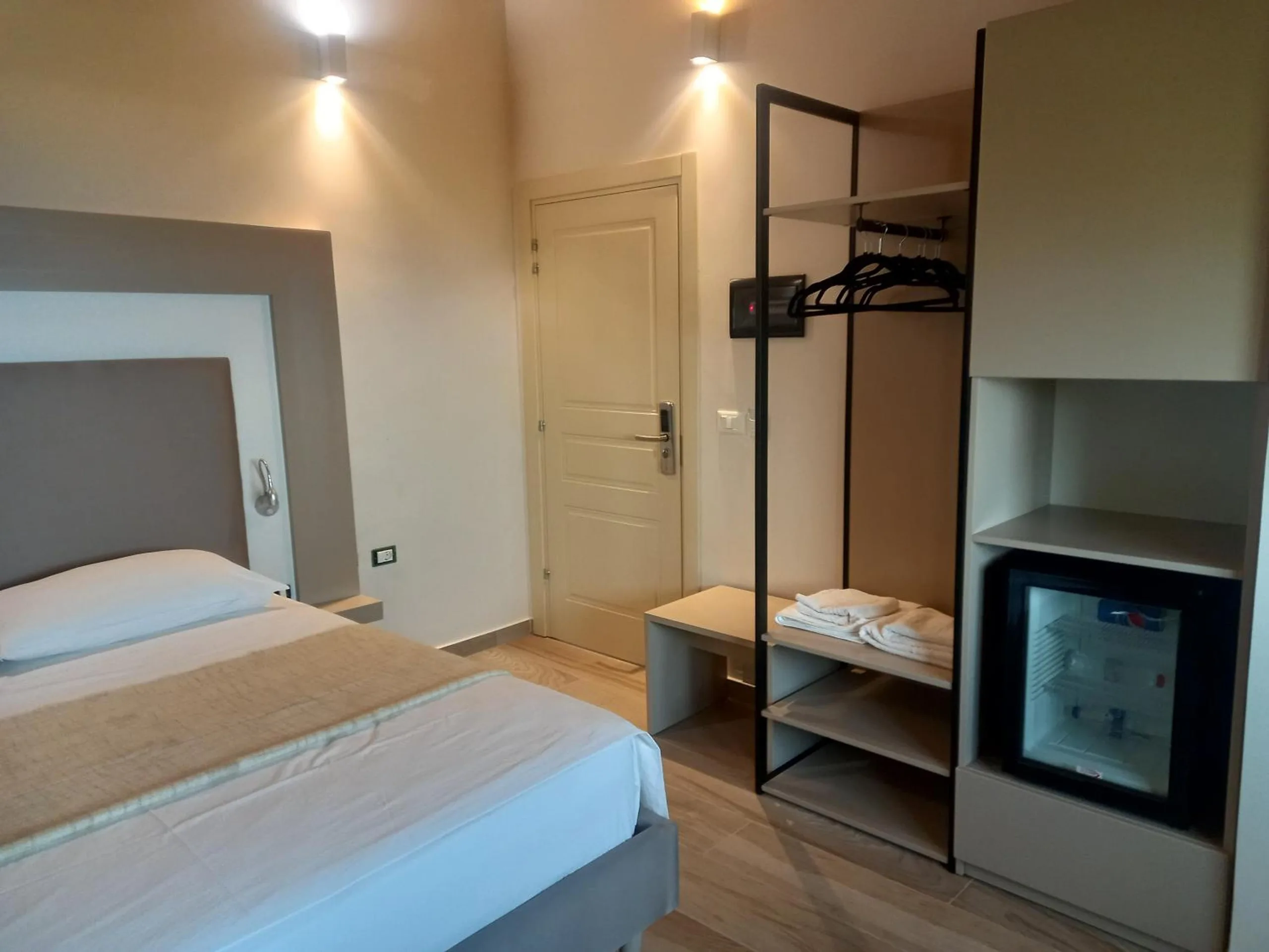 A double bed beside an open black metal wardrobe with towels and a small glass-fronted fridge