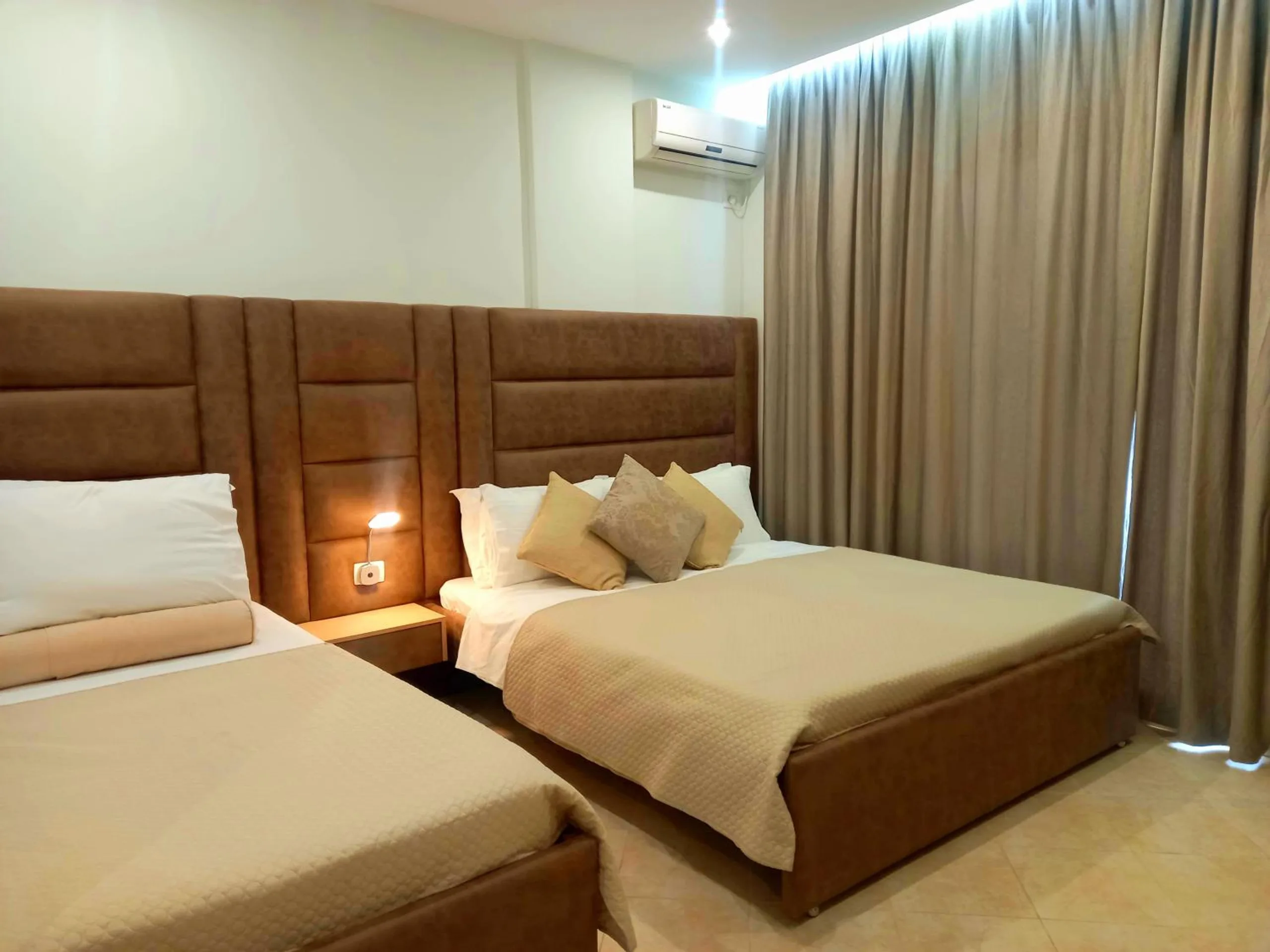 A double bed and a single bed side by side, both with tall tan headboards, in a family room with cream tiles