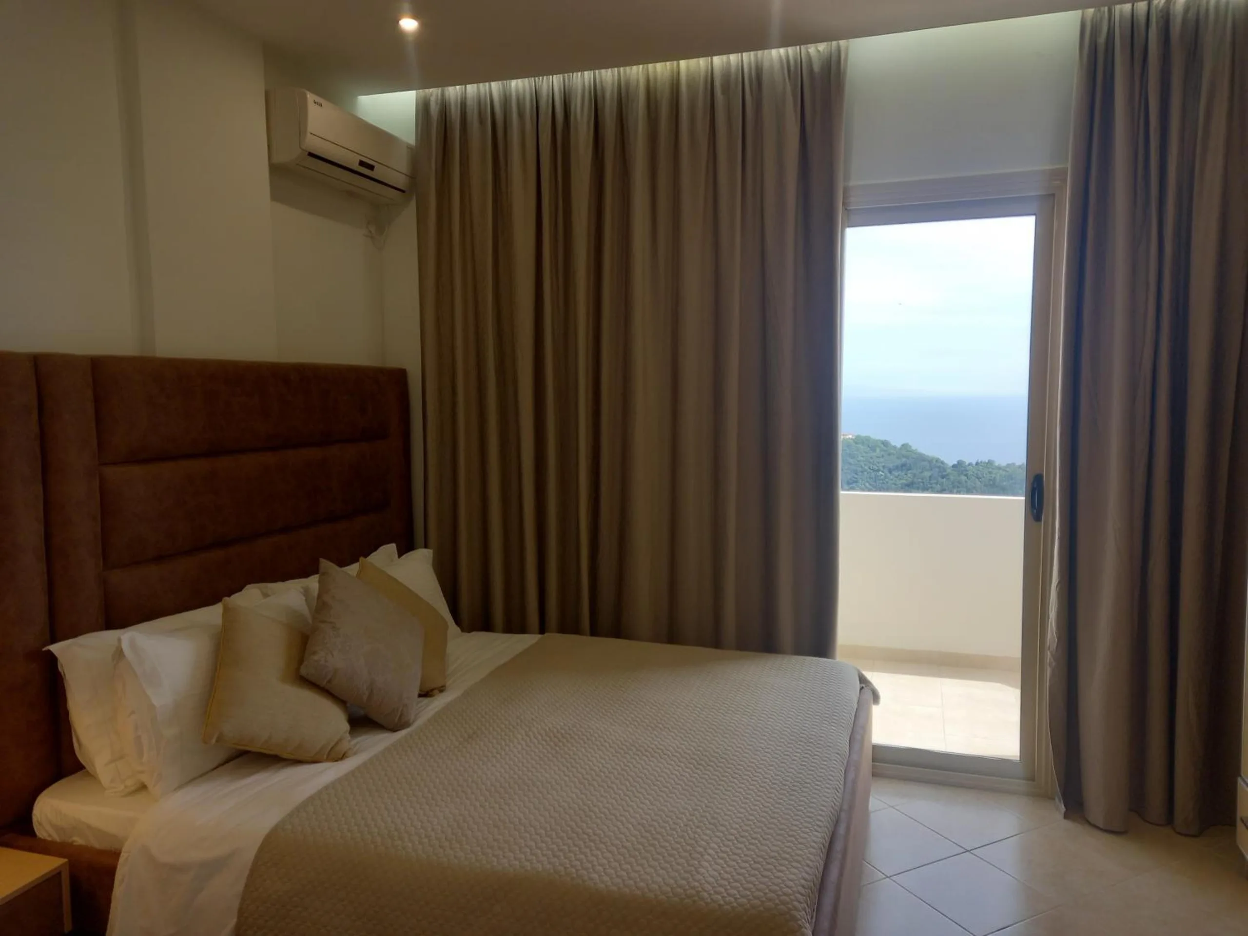 A double bed with a tall tan headboard and long curtains drawn back from a balcony door with the sea beyond the hillside