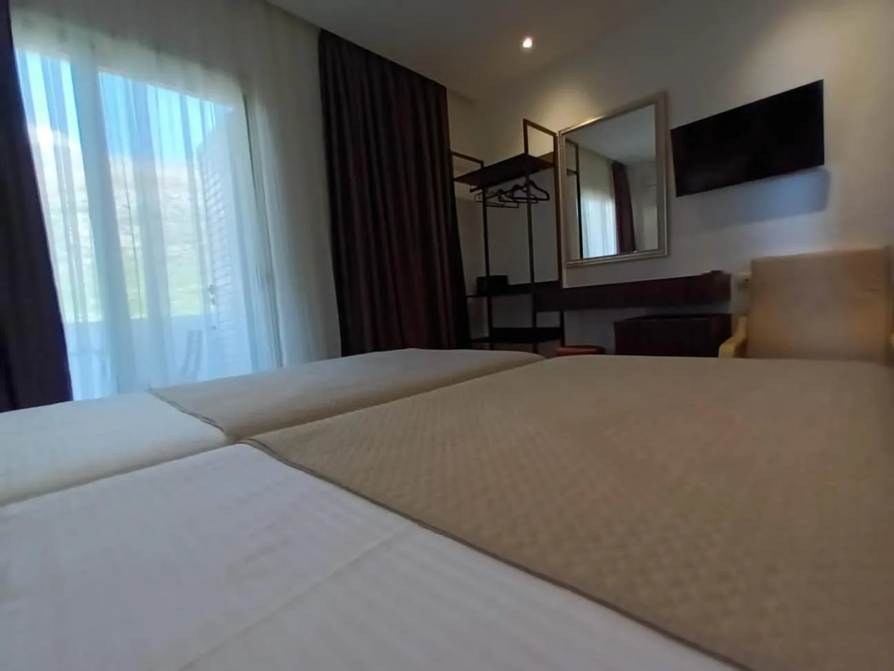 Two single beds pushed together with a beige quilted cover, white curtains drawn over a bright window