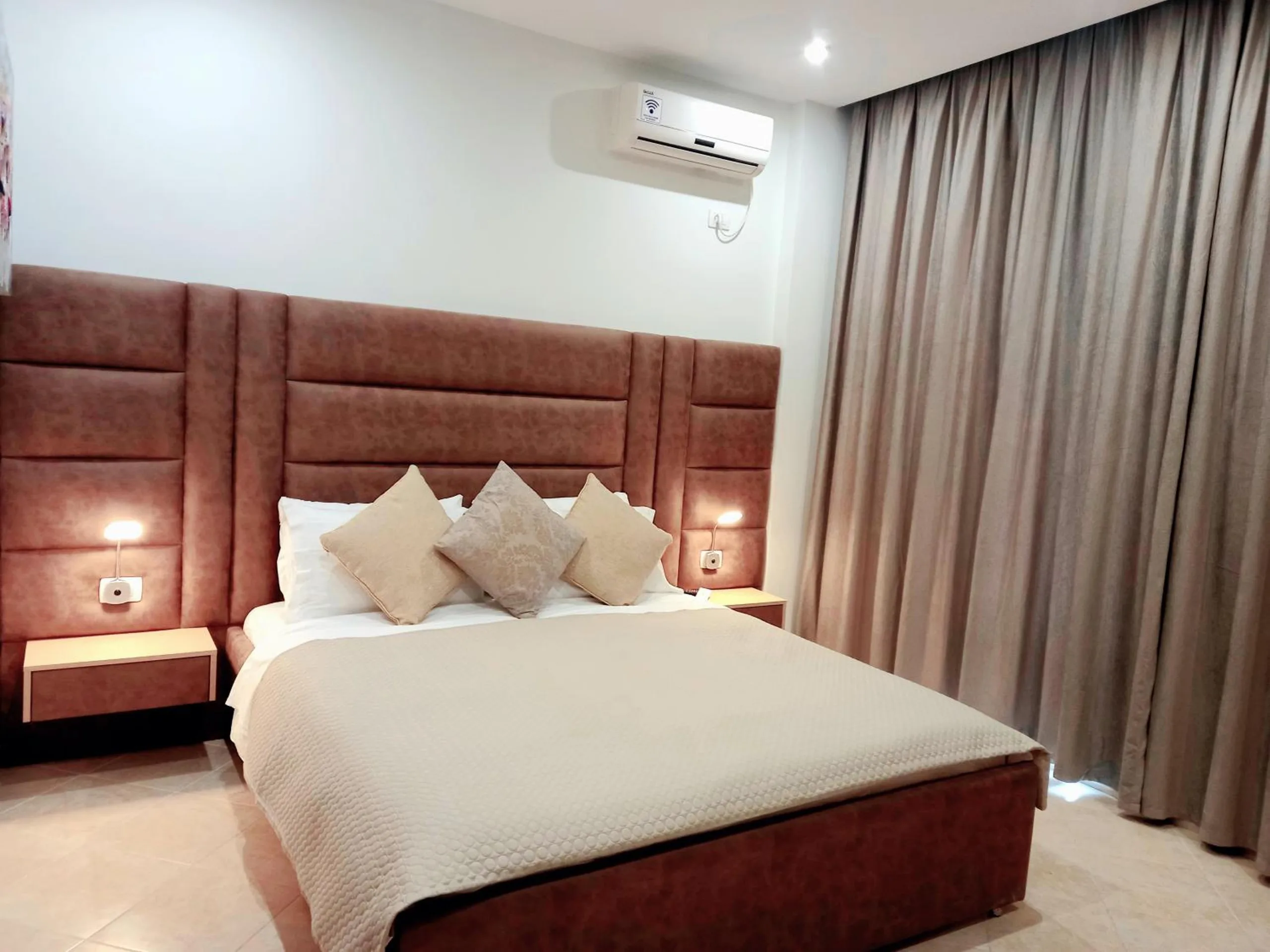 A double bed with a tall tan headboard and cushions, lit by two bedside lamps