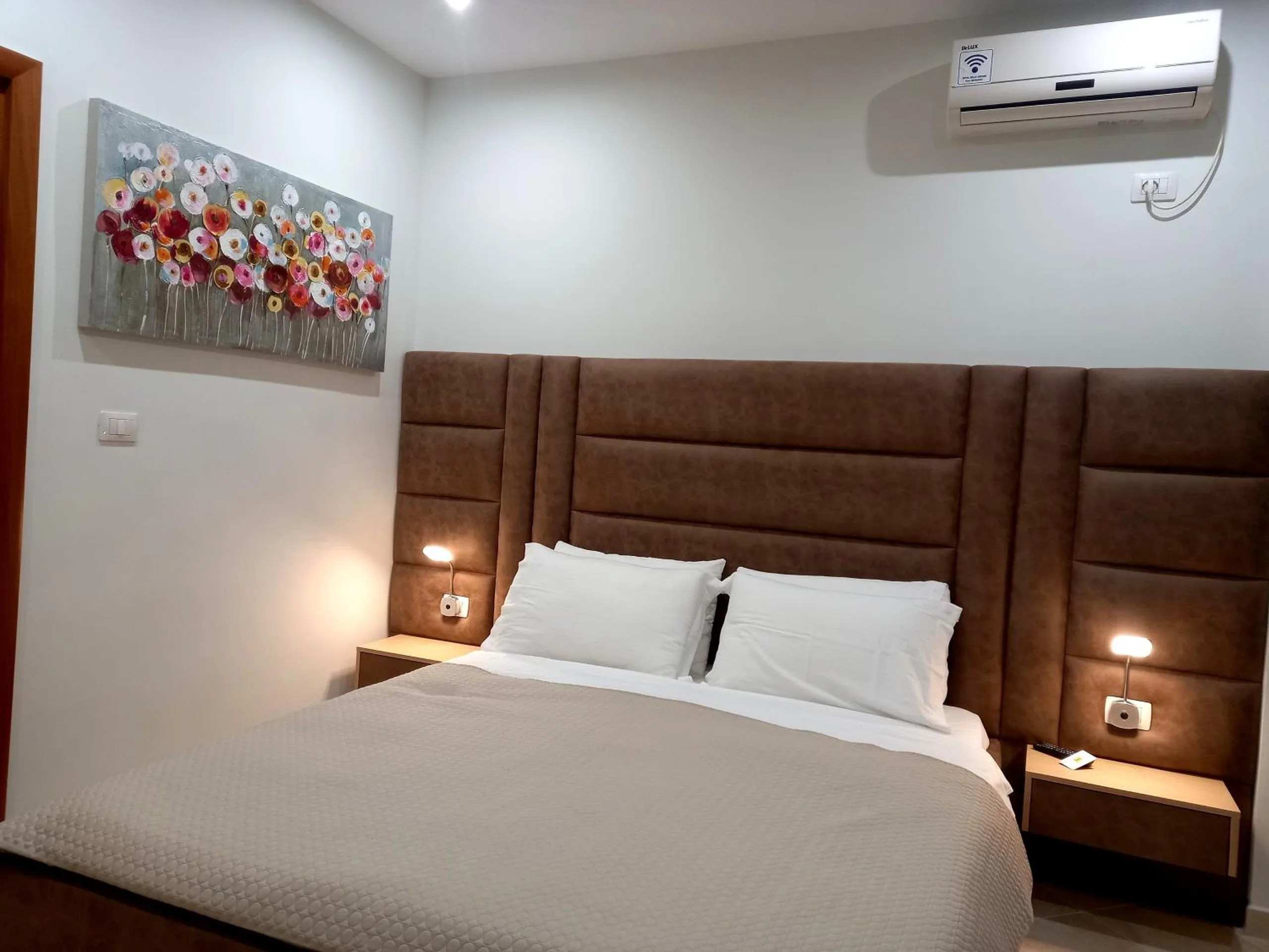 A double bed with a tall tan upholstered headboard and white pillows under a flower painting